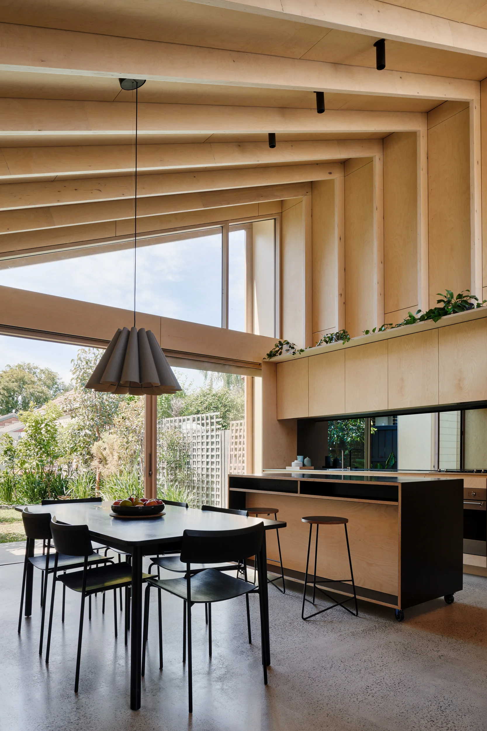 A modern dining space features plywood paneled walls, a vaulted ceiling, a black dining table with chairs, and sliding glass doors opening to a garden.