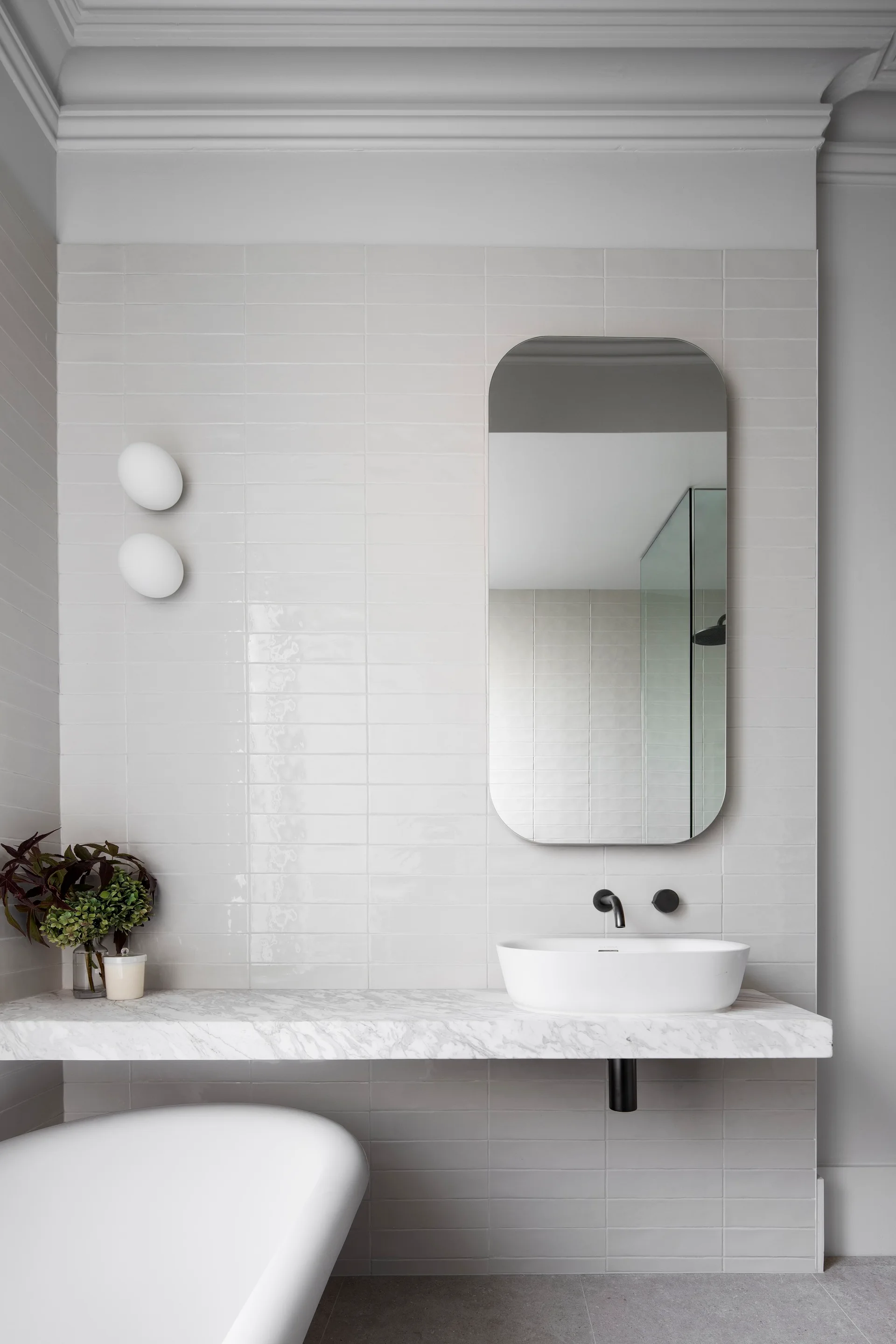 A bathroom vanity features a floating marble countertop, vessel sink, and an oblong mirror against a light grey tiled wall.