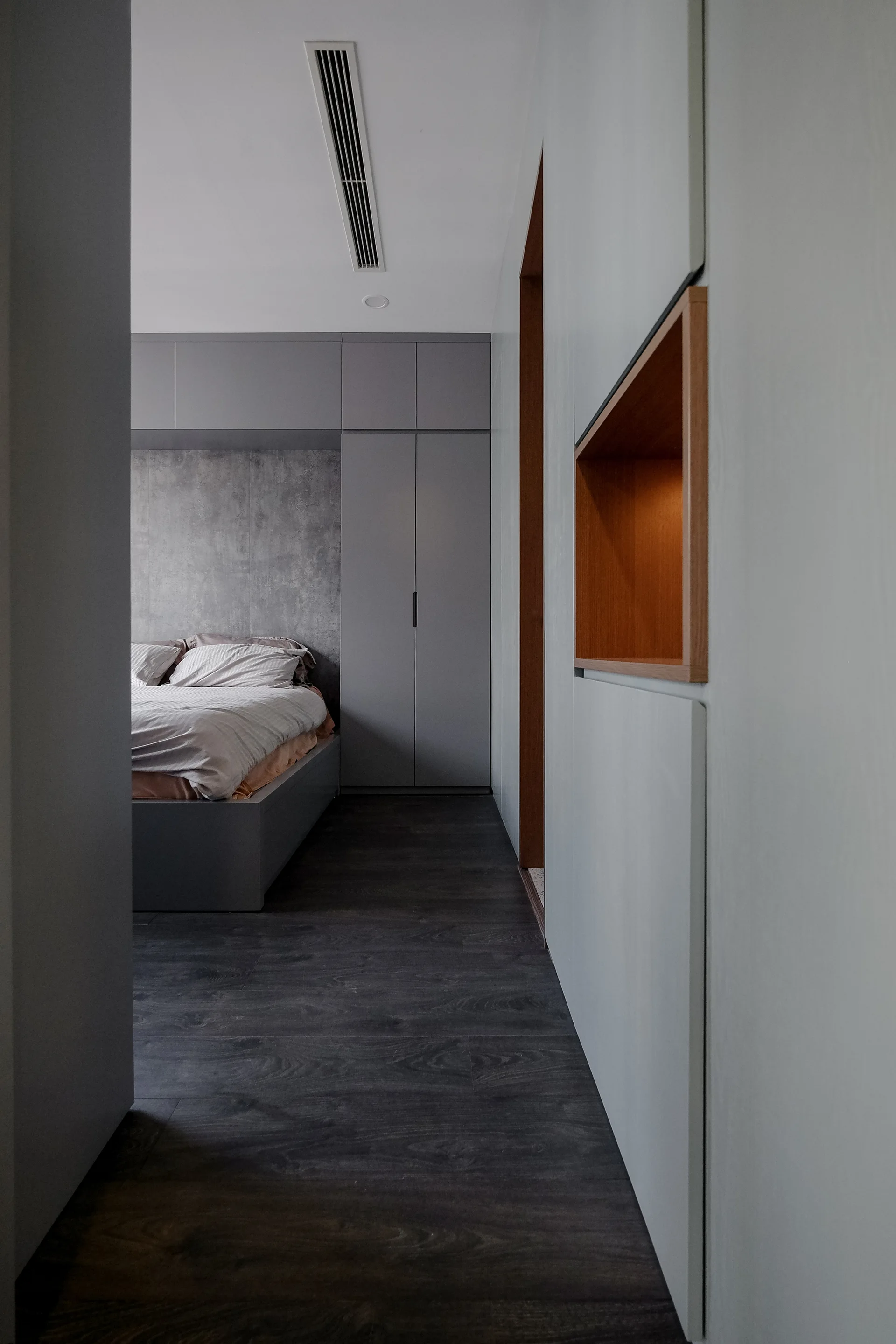 A minimalist bedroom features custom matte gray wardrobes framing a platform bed, seen past a wall with a recessed warm wood niche.