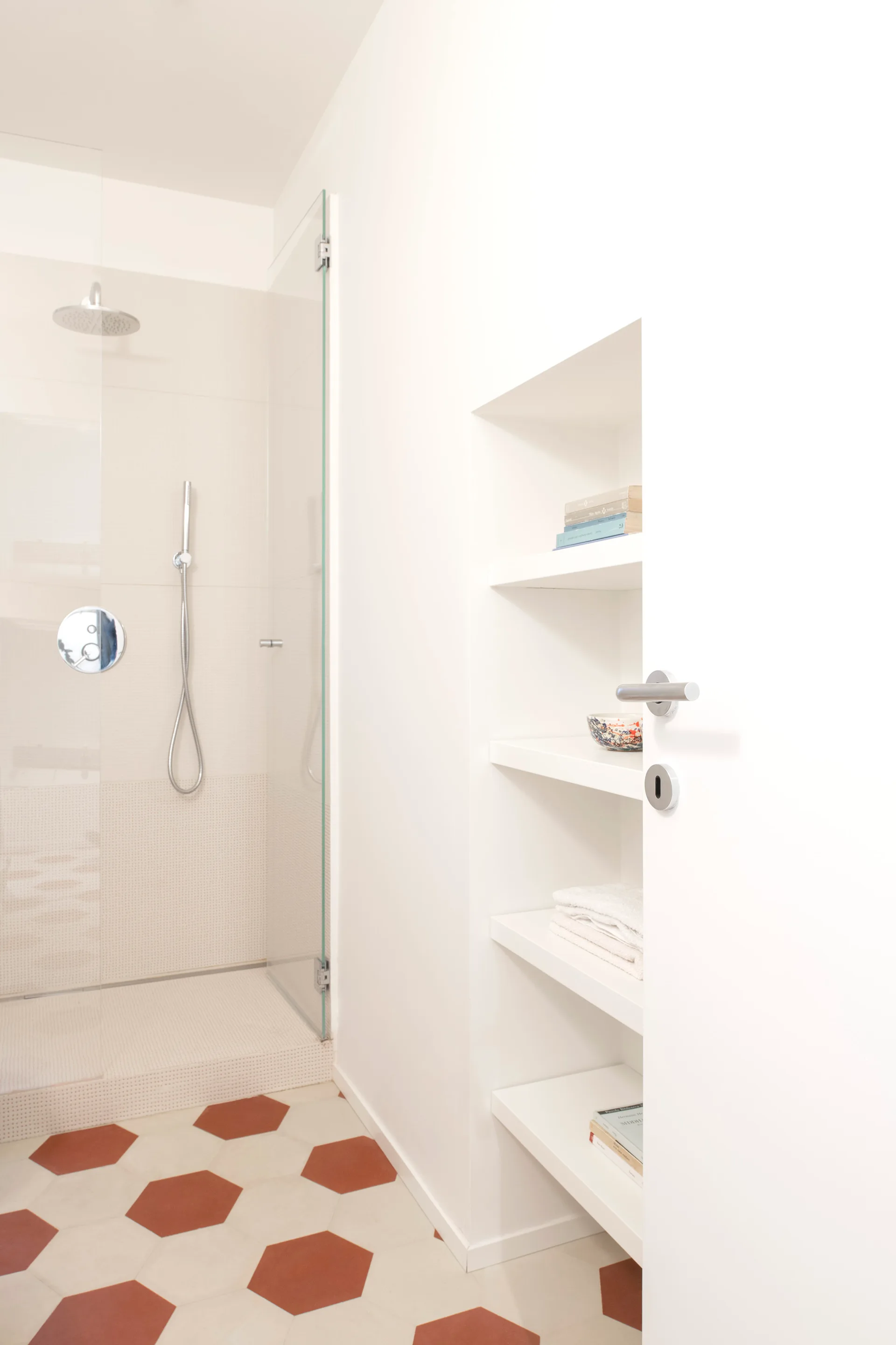 A modern white bathroom featuring terracotta and white hexagonal floor tiles, a glass walk-in shower, and a built-in recessed shelving niche.