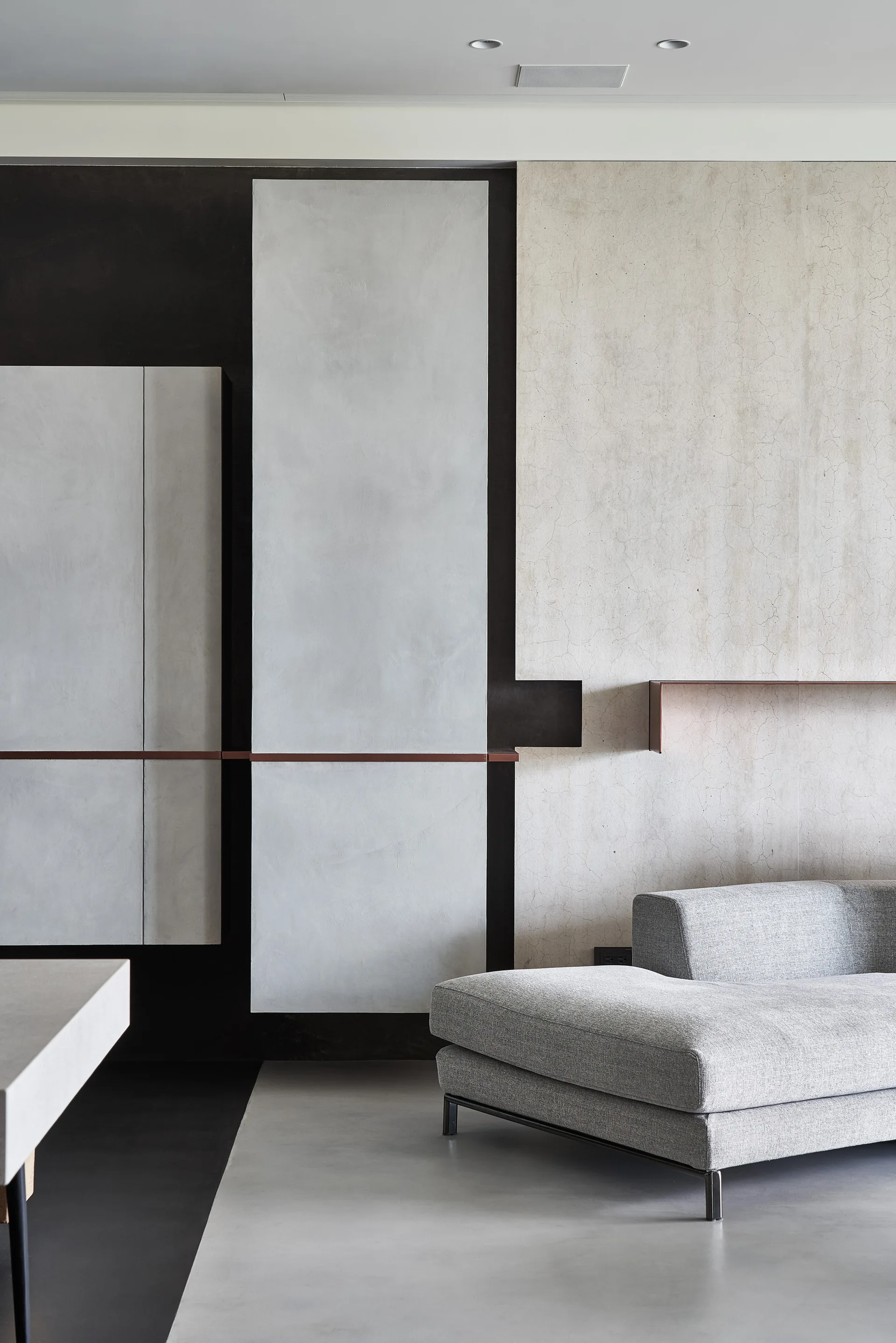 A low-profile grey sofa sits against a wall made of geometric plaster panels in charcoal, light grey, and cream with a thin copper accent shelf.