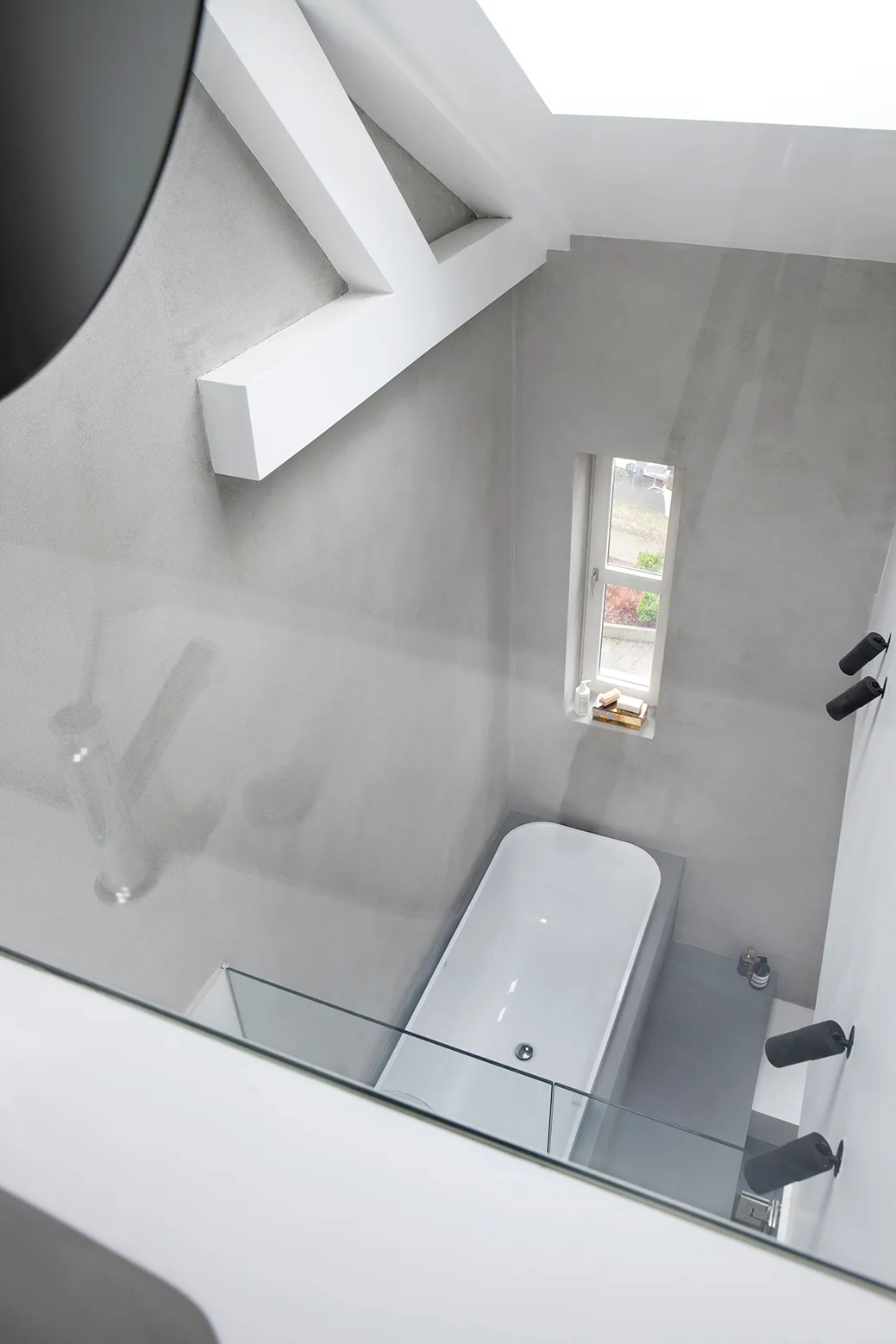 A high-angle view looking down at a white bathtub surrounded by matte grey concrete walls, featuring a narrow vertical window and white ceiling beams.