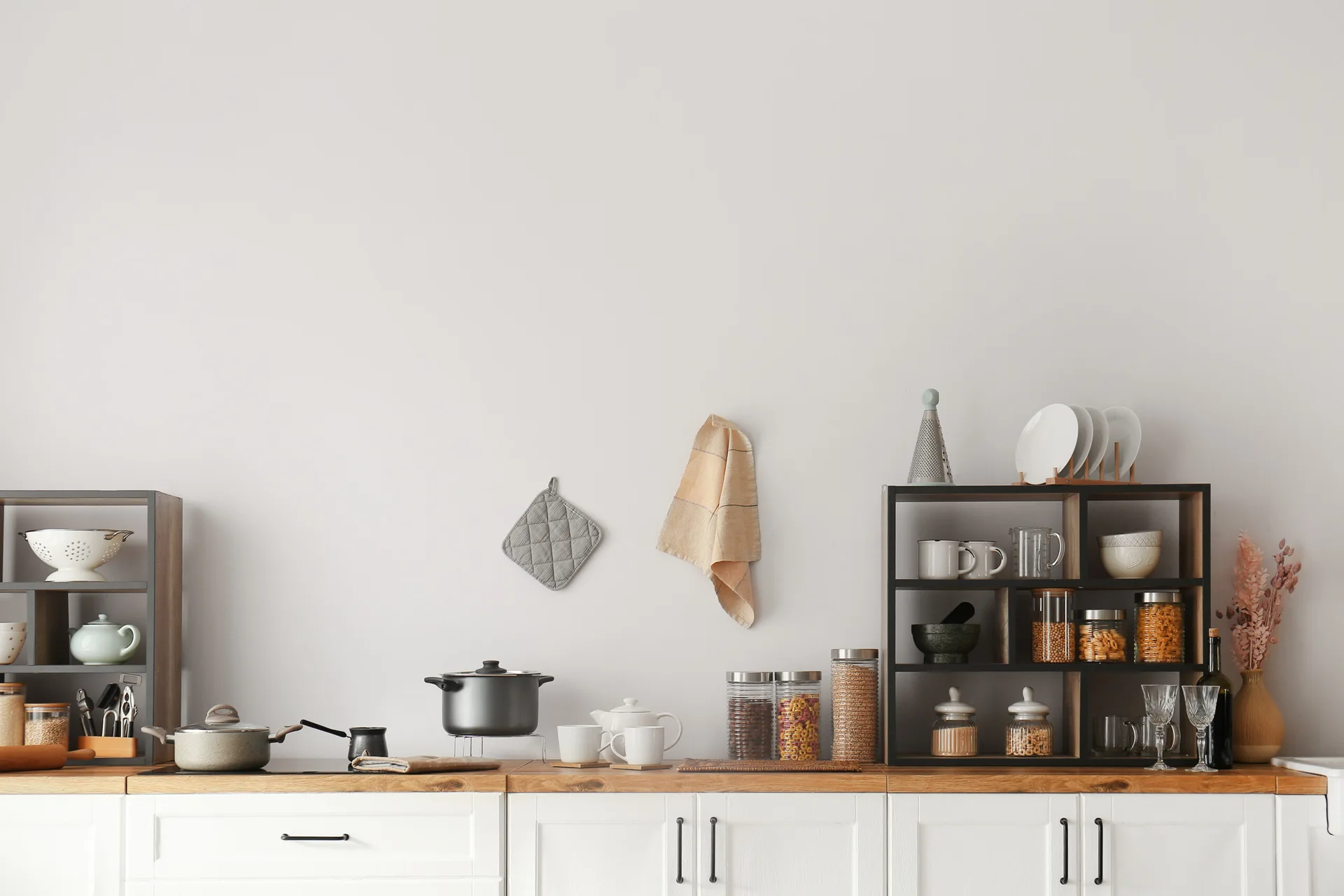 White shaker cabinets with a wood countertop feature black open storage shelves filled with jars, cups, and kitchen accessories against a plain wall.