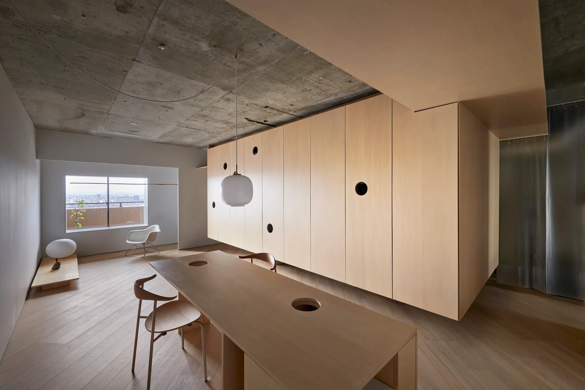 A minimalist dining room features a long wooden table, a suspended light wood storage unit with circular cutouts, and an exposed concrete ceiling.
