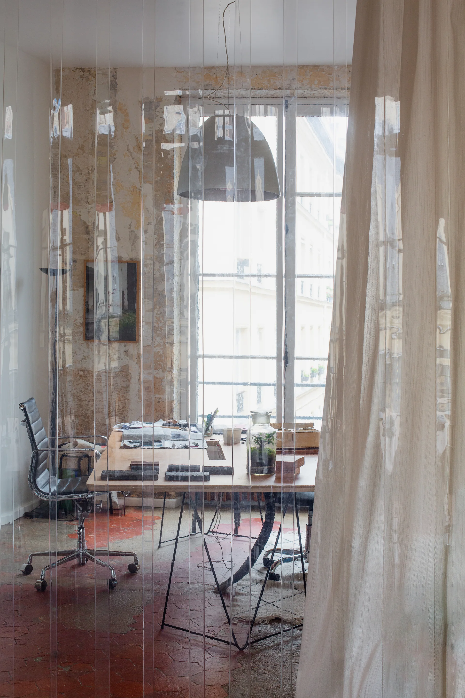 An industrial office workspace viewed through a clear plastic strip curtain, featuring a wooden desk, task chair, and distressed plaster walls.