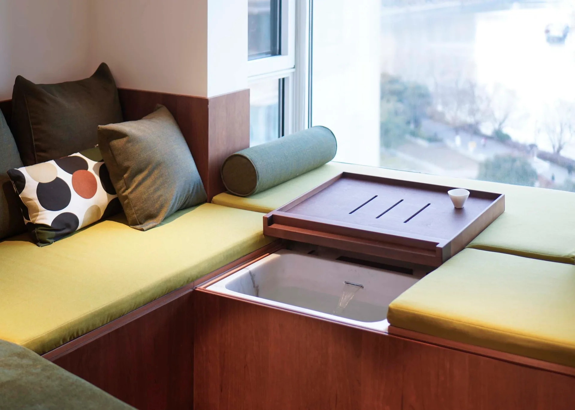 A built-in wooden window seat features sage green cushions, a small basin with running water, and a removable wooden tray.