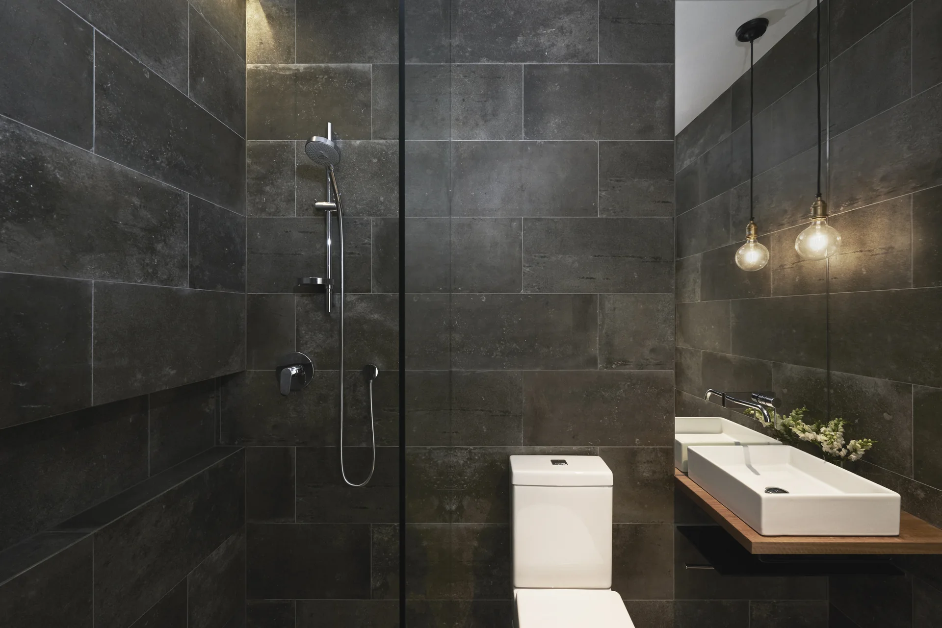 A minimalist bathroom features charcoal stone tile walls, a white toilet, and a vessel sink resting on a floating wood shelf.