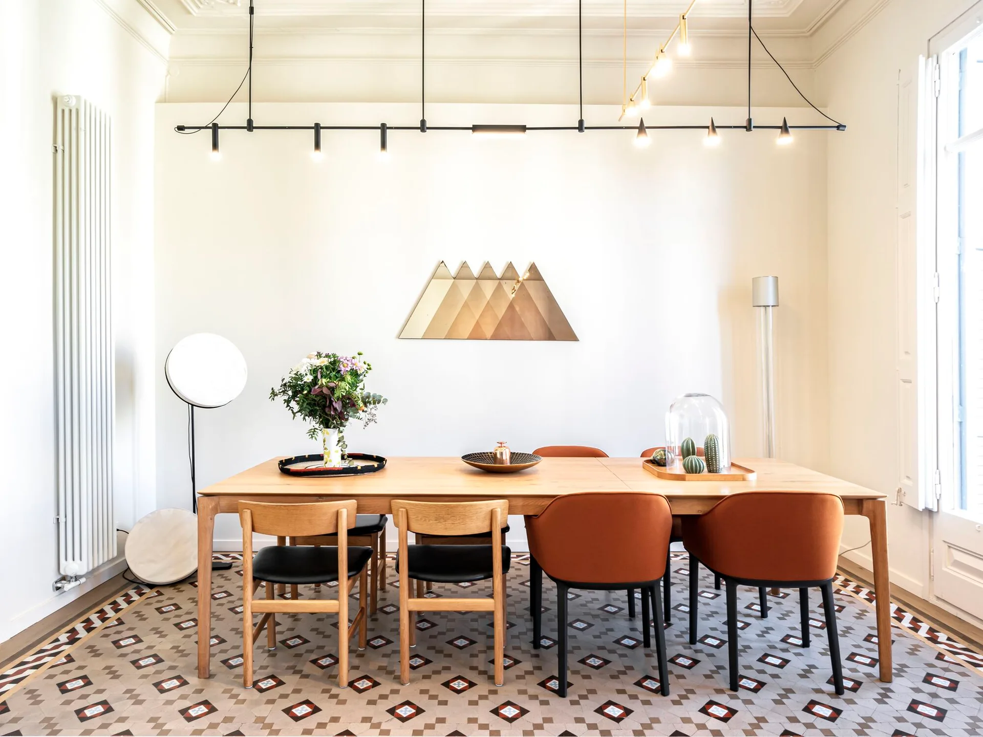 A light wood dining table with orange and wood chairs sits on a geometric tiled floor beneath a black linear pendant light.