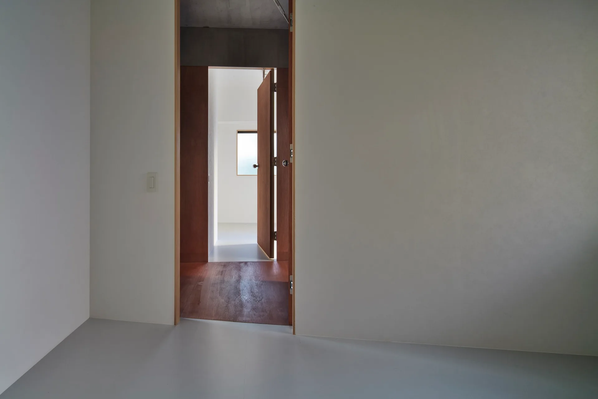 An open wood door and warm plywood flooring create a transition zone between white minimalist rooms under an exposed concrete ceiling.