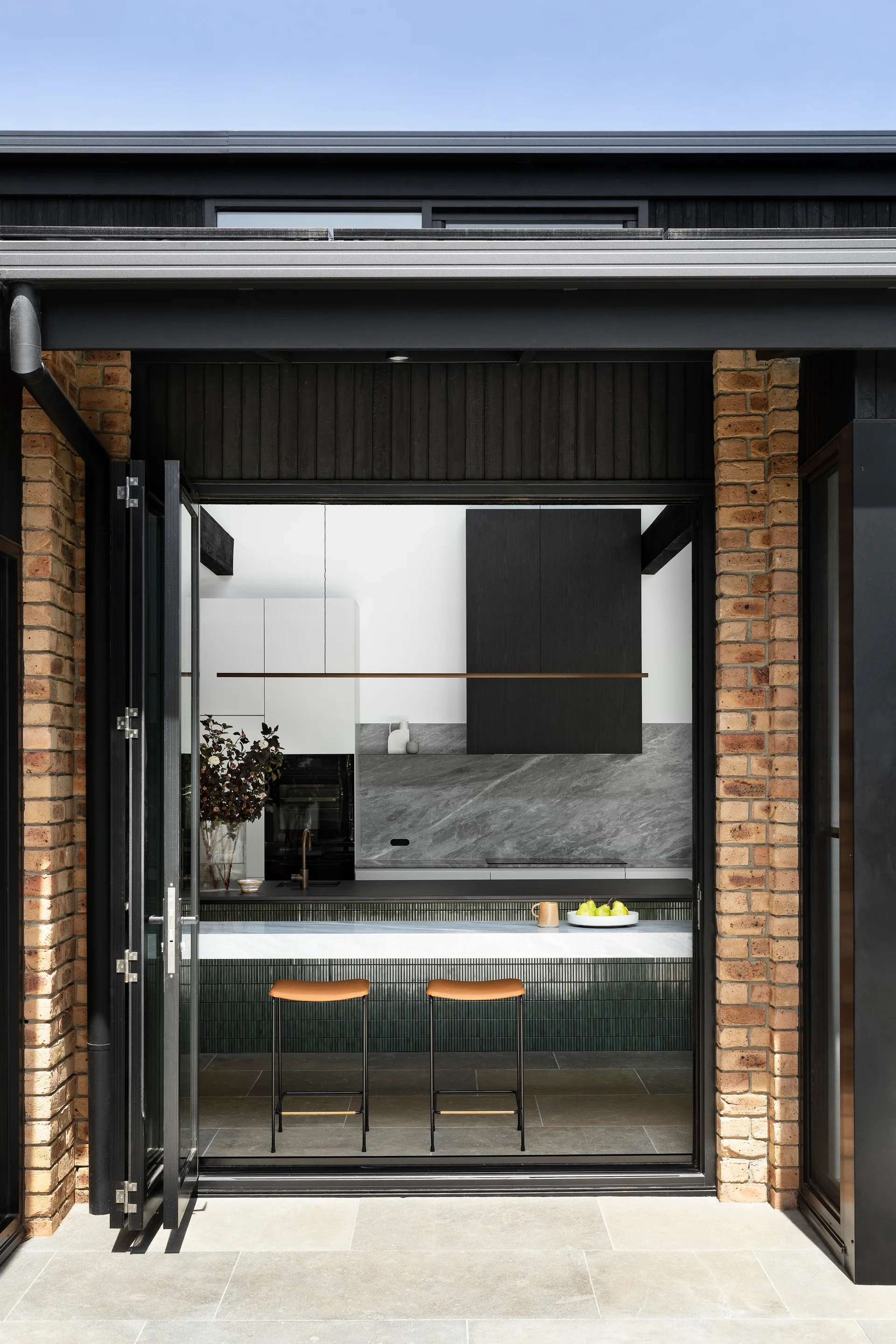 A view through open folding doors framed by brick pillars reveals a contemporary kitchen with a veined stone backsplash and a stone-topped island.