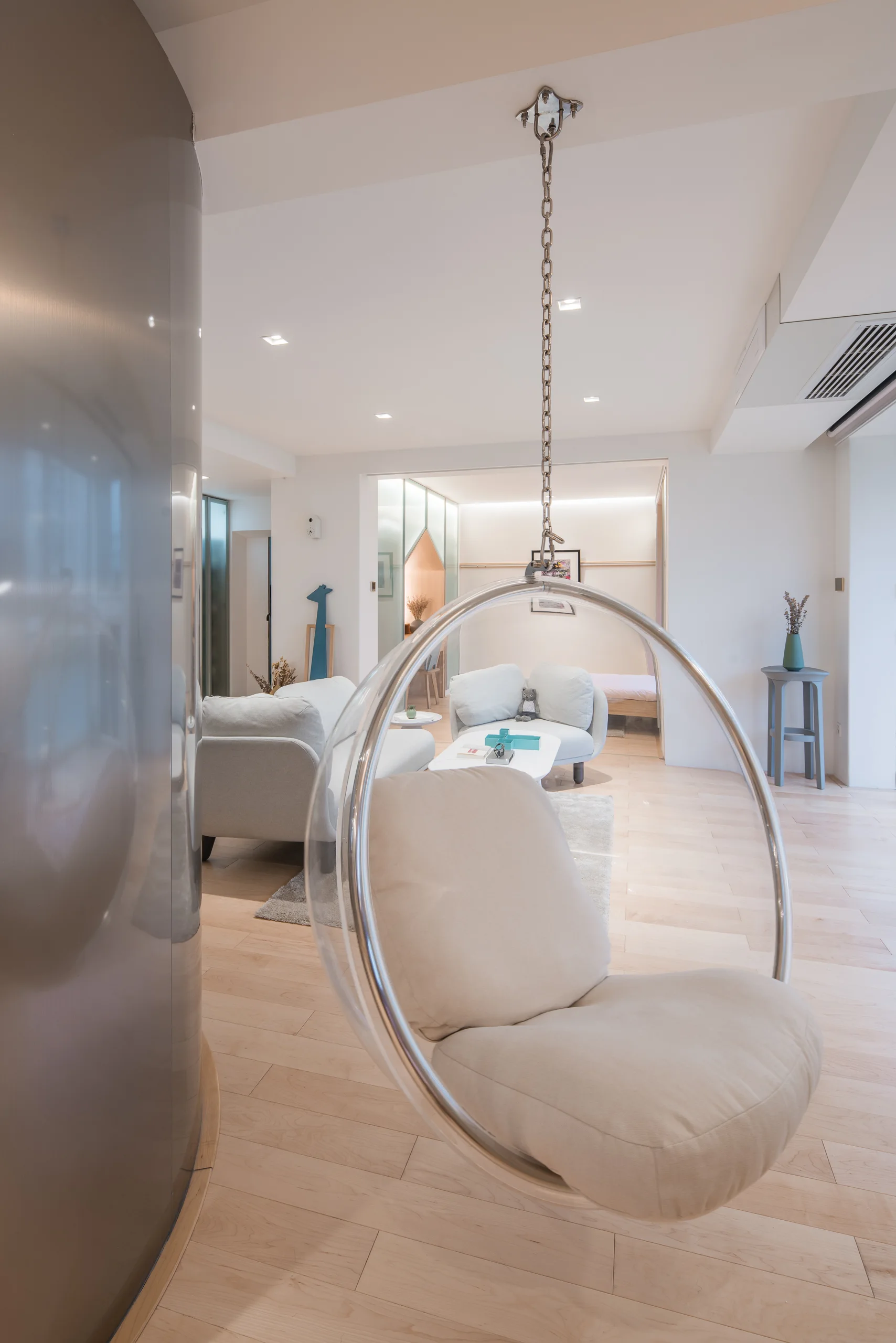 A clear acrylic bubble chair hangs from the ceiling in a minimalist living room with a light grey sofa and blonde wood floors.