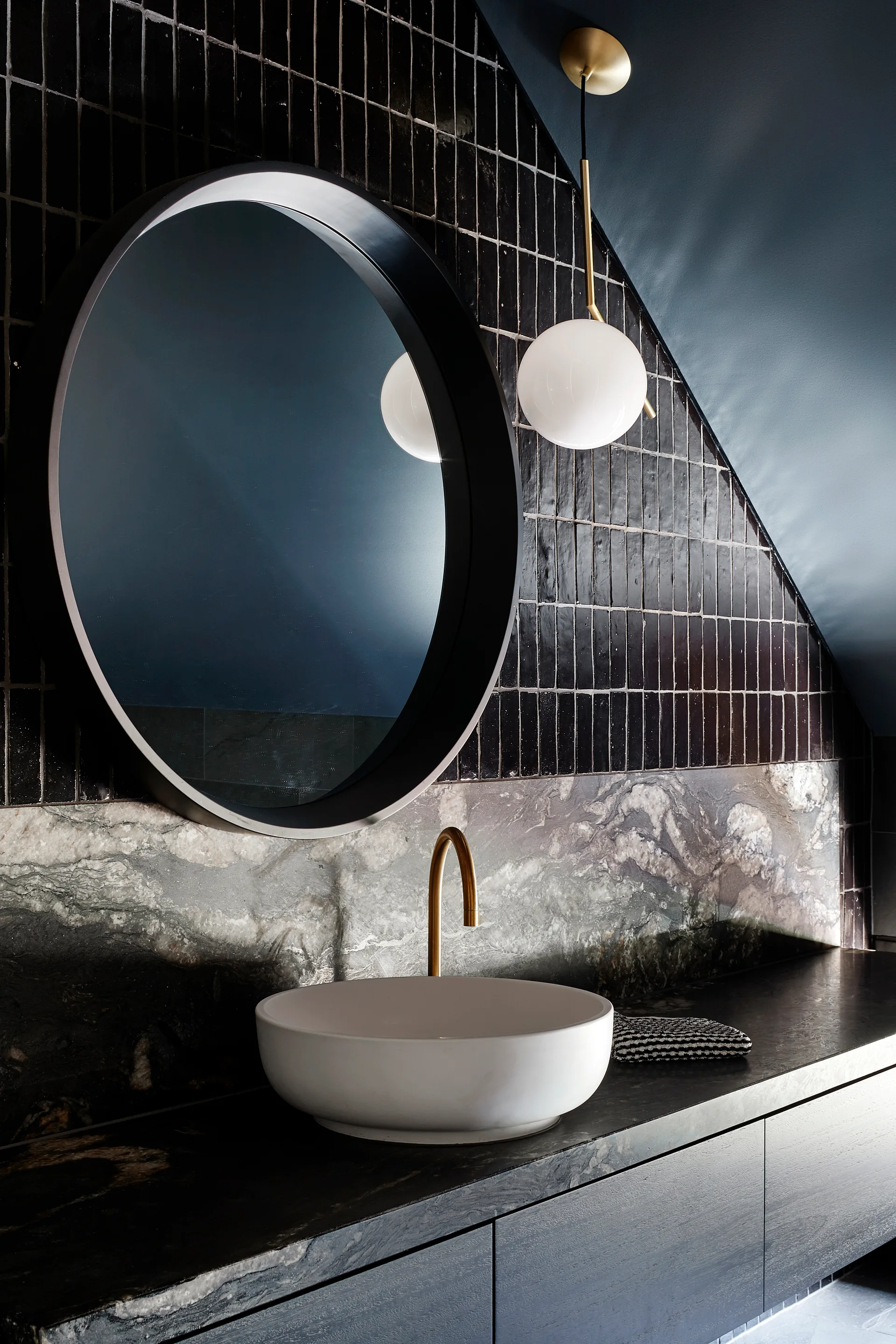 A white vessel sink sits on a dark vanity below a round black mirror, backed by glossy black tiles and a veined stone backsplash.