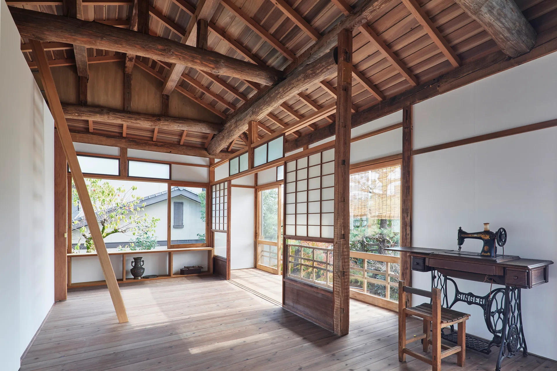 A bright Japanese interior features an exposed timber ceiling structure, sliding shoji screens, wood flooring, and a vintage treadle sewing machine.