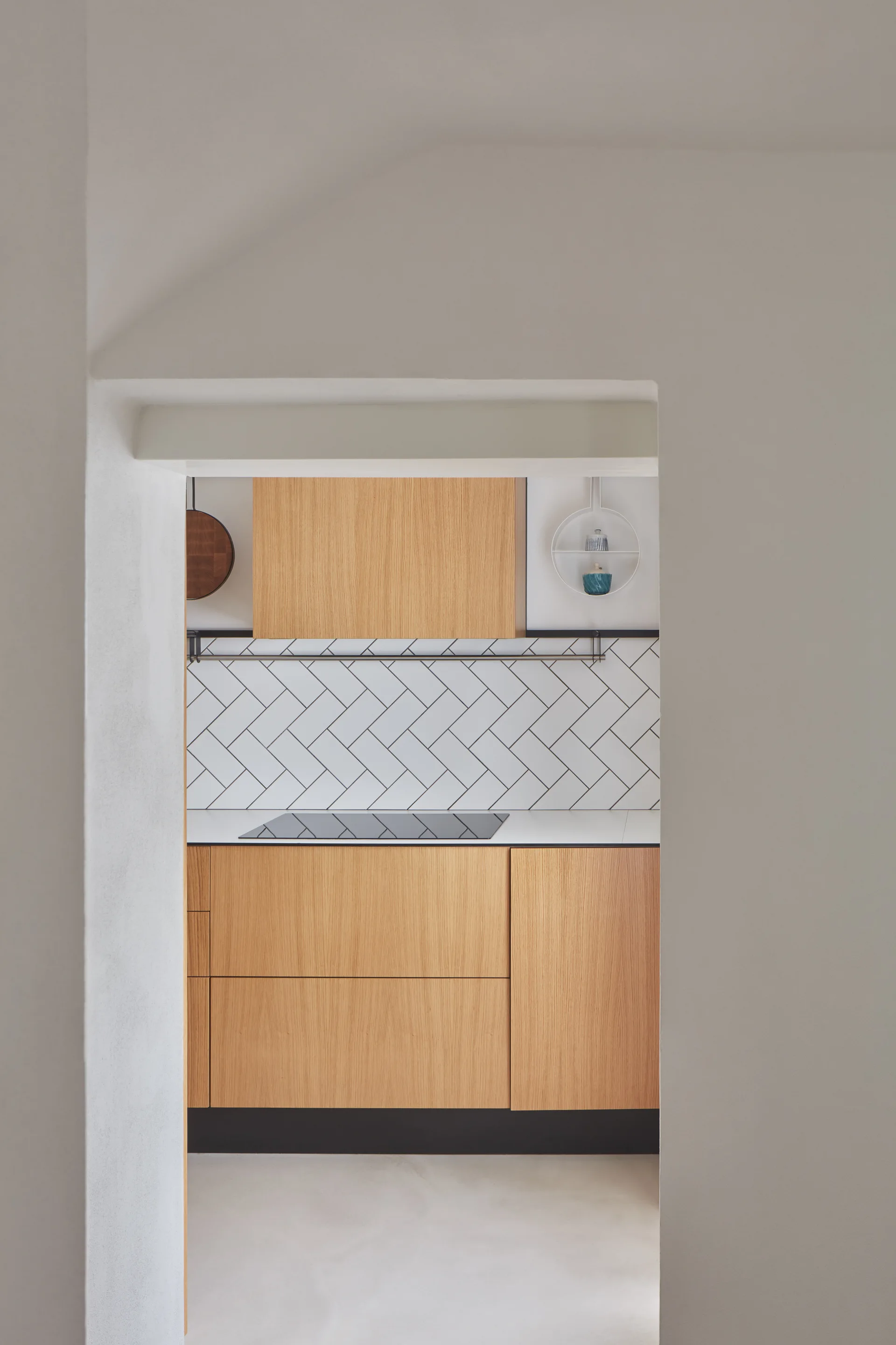A view through a plaster doorway showing a modern kitchen with flat-panel wood cabinets, a white herringbone tile backsplash, and an induction cooktop.