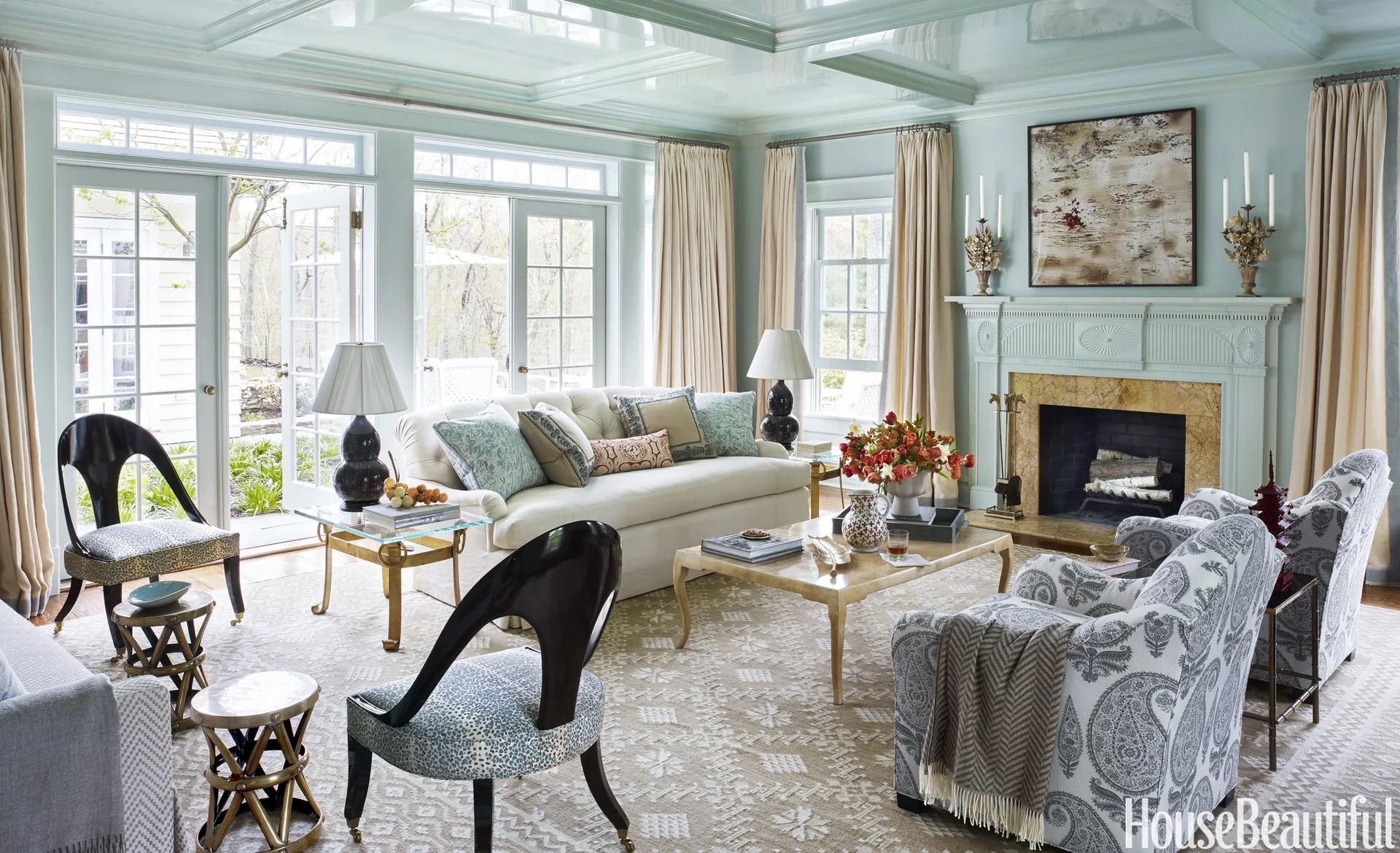 A pale blue living room features a cream tufted sofa, patterned armchairs, and a fireplace beneath a glossy coffered ceiling.