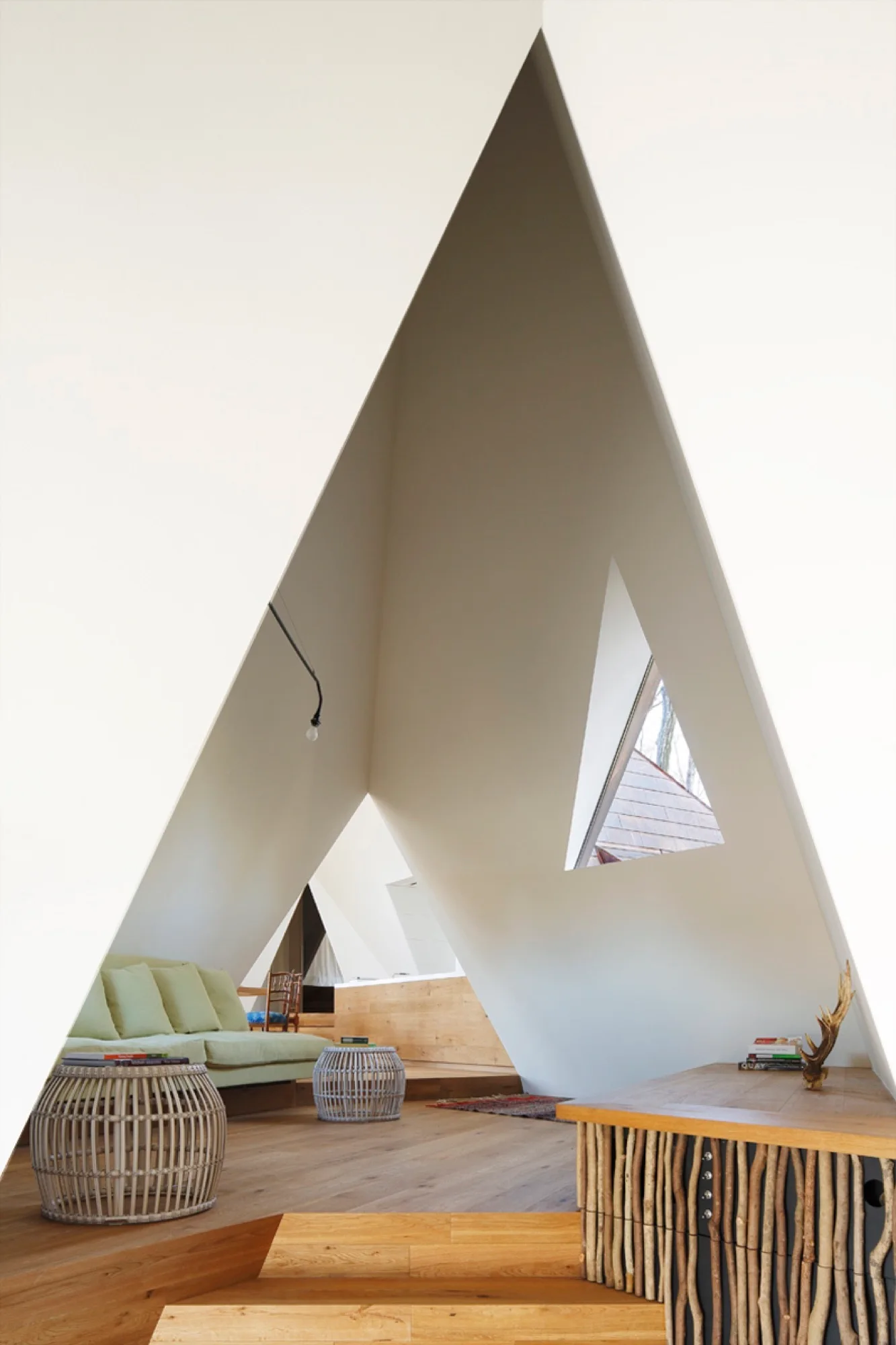 An interior view of a minimalist room featuring tall, angled white walls that form a triangular peak over wood floors and light green seating.
