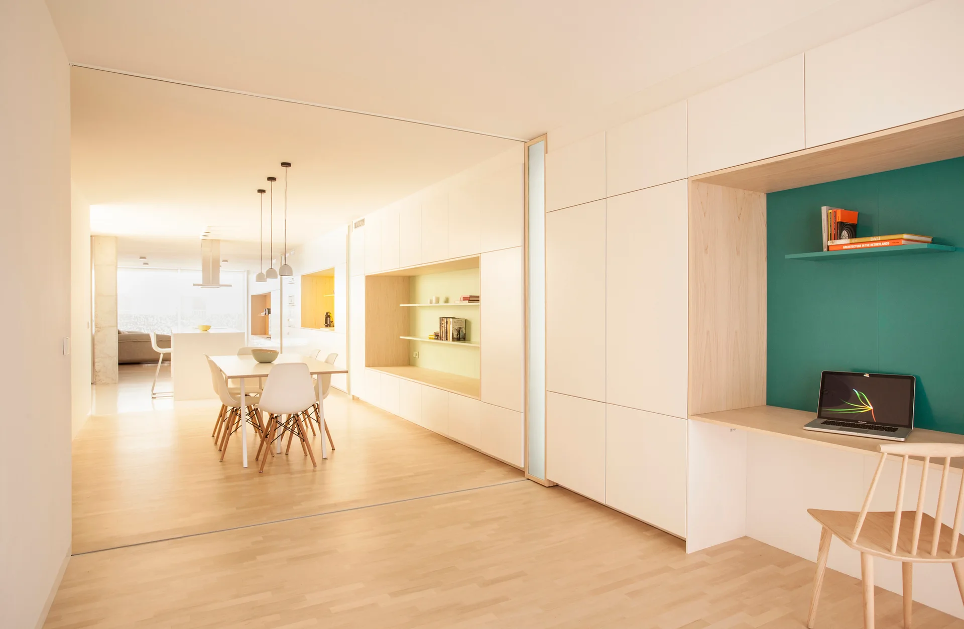 A minimalist room features a white built-in wall cabinet with an integrated light wood desk, a teal accent panel, and an adjacent dining area.