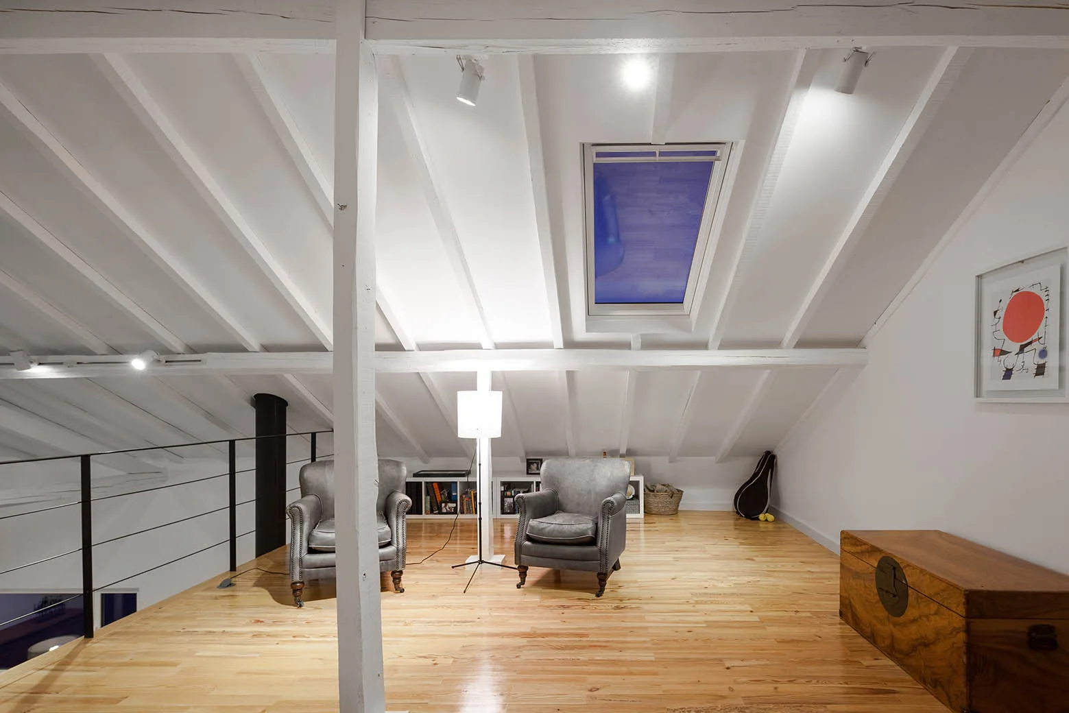 A loft attic space featuring two gray leather armchairs on glossy wood floors beneath a slanted white ceiling with exposed beams and a skylight.