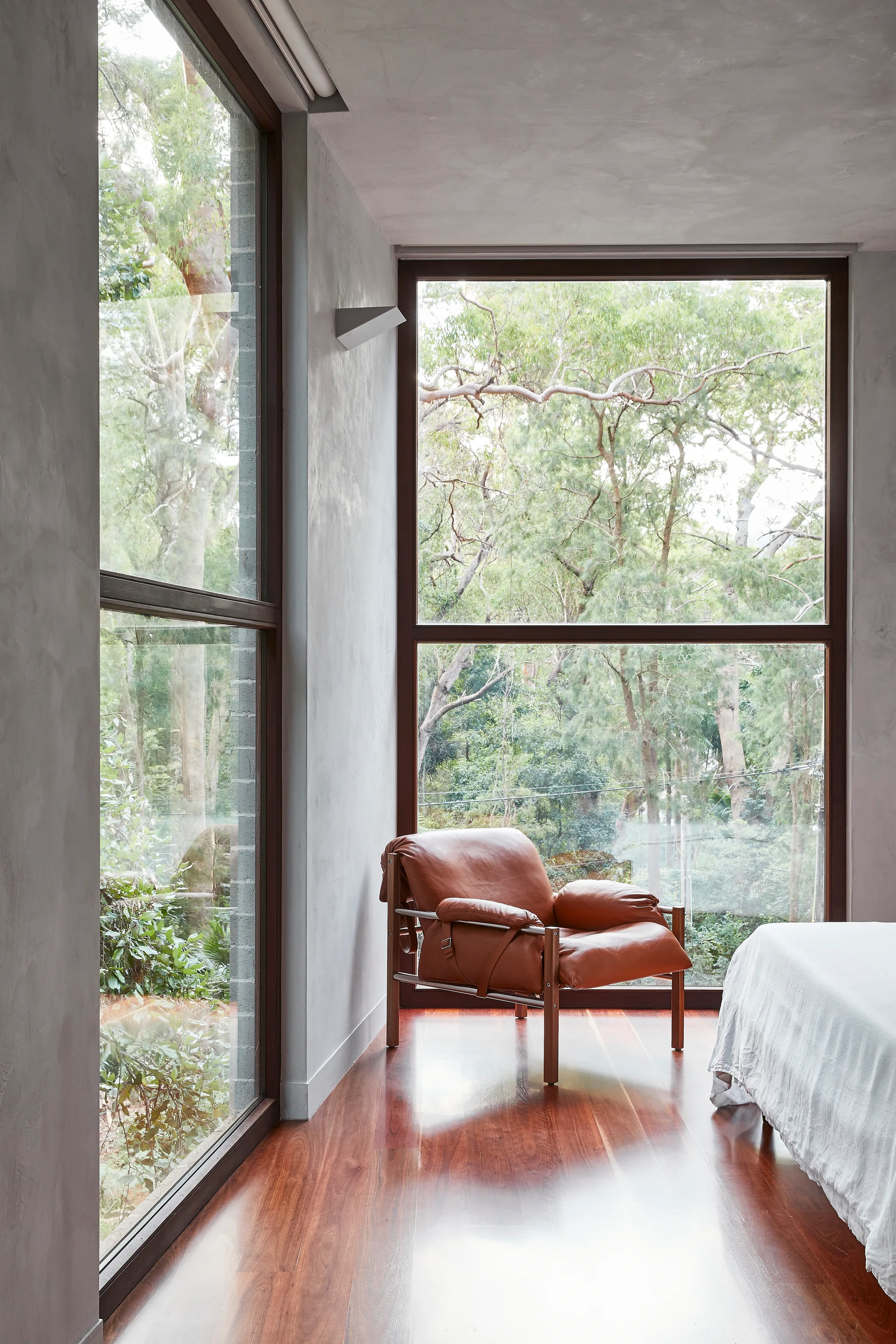 A brown leather lounge chair sits on polished wood floors in a grey plaster bedroom corner beside large windows looking out at trees.