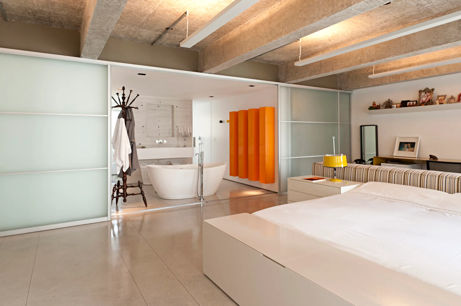 A contemporary bedroom with a white bed and exposed concrete ceiling beams connects to an ensuite bathroom through frosted glass sliding doors.