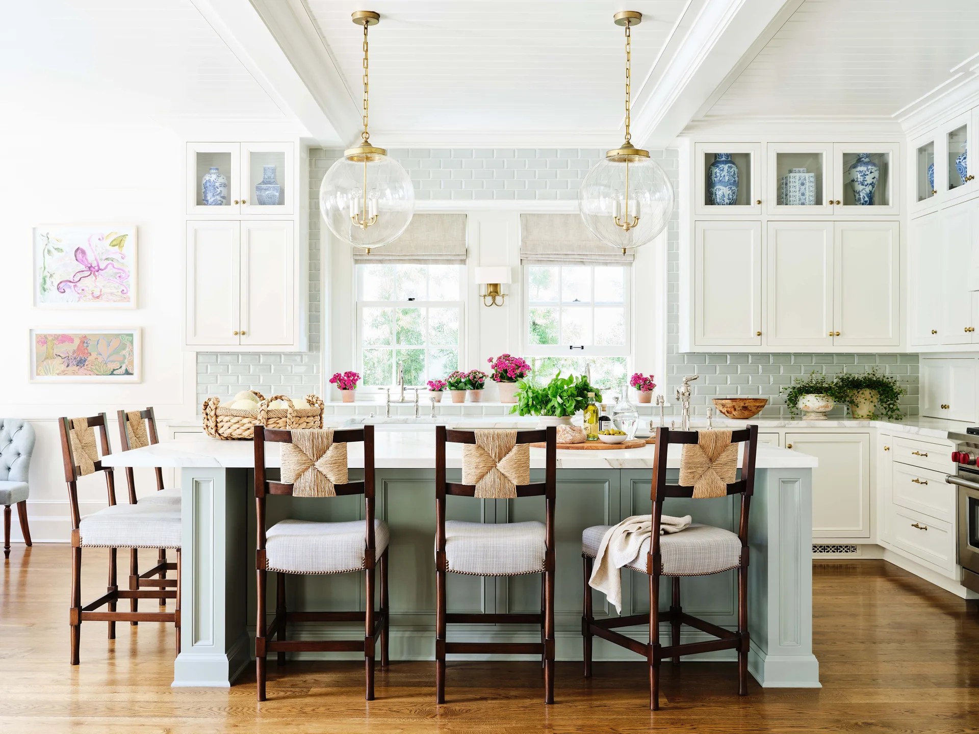 A bright kitchen featuring a sage green island with woven back stools, white cabinetry, a soft green subway tile backsplash, and glass globe pendants.