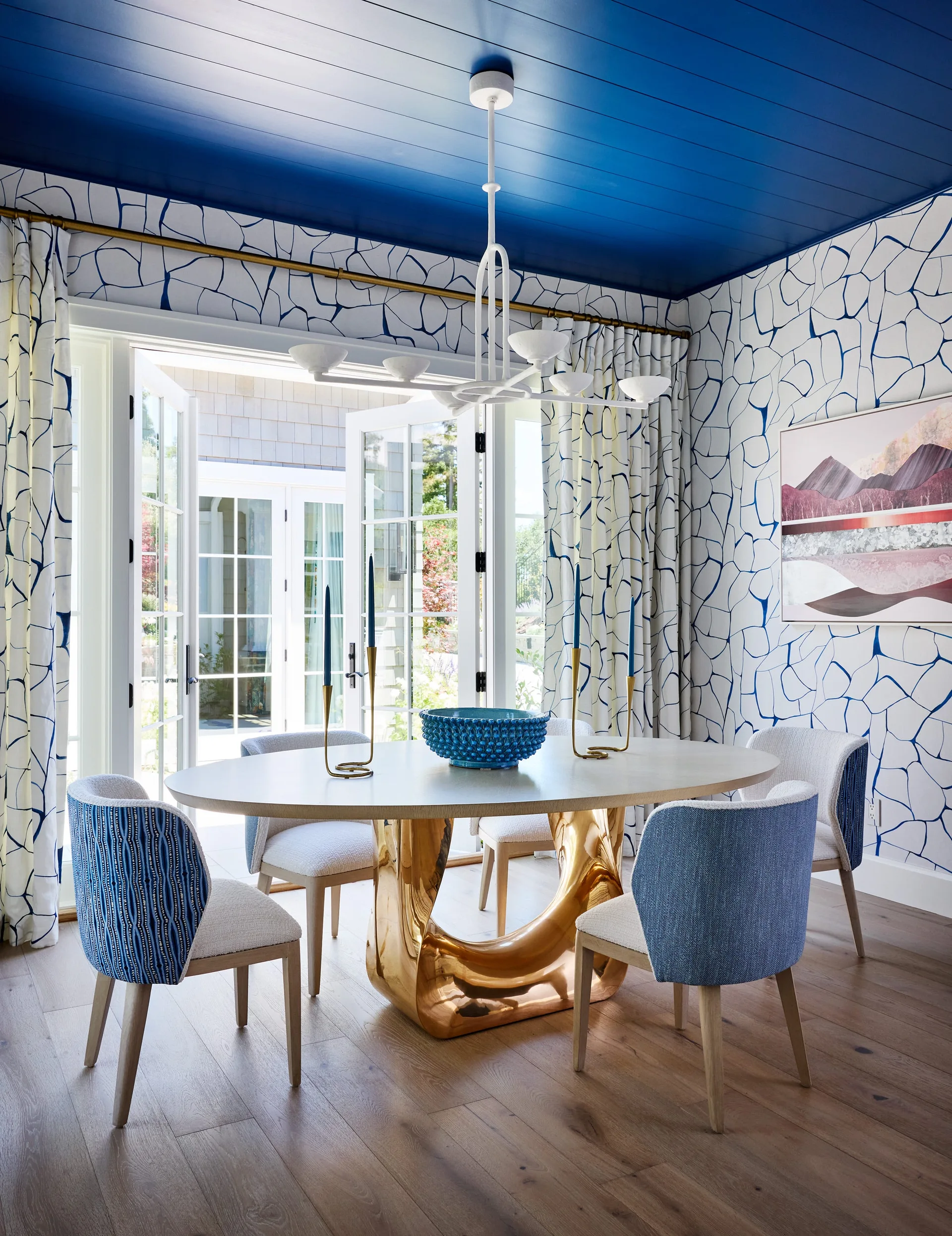 A dining room featuring a blue paneled ceiling, blue and white graphic wallpaper, and an oval dining table with a sculptural gold base.