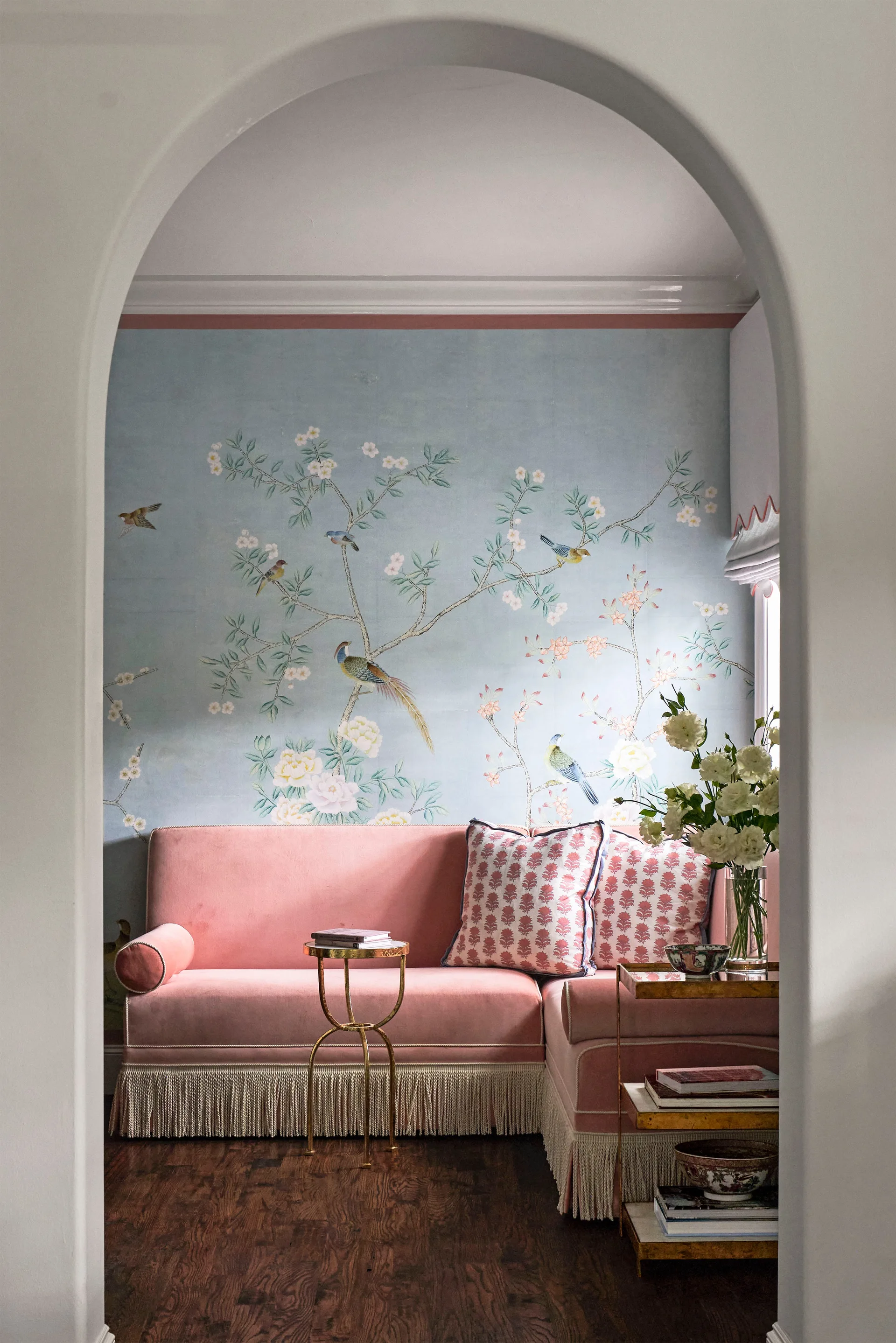 A pink velvet sofa with fringe trim sits against a light blue floral wallpaper background, viewed through a white arched doorway.