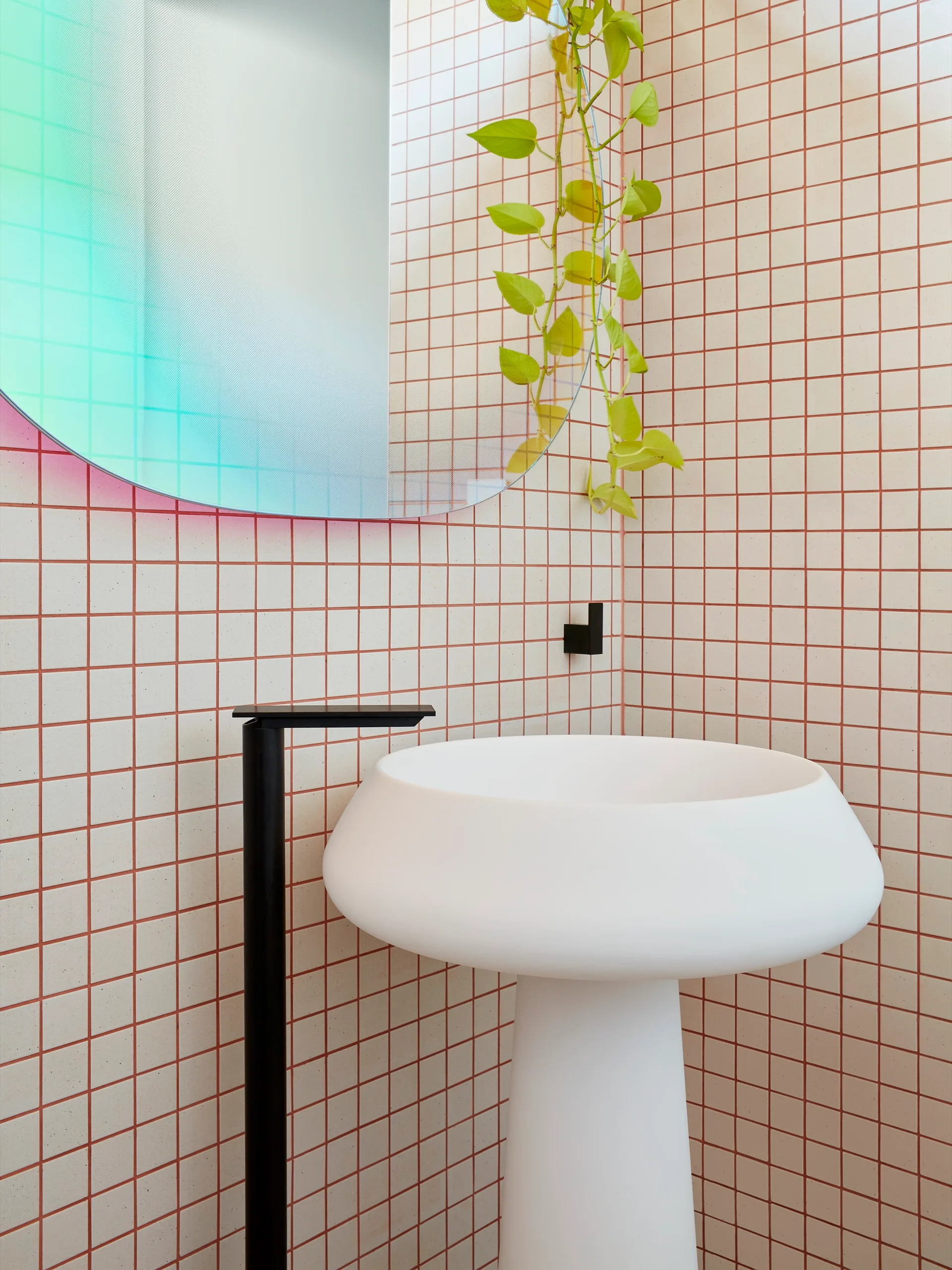 A modern white pedestal sink with a tall black faucet stands against a cream tiled wall with red grout lines next to a round mirror.