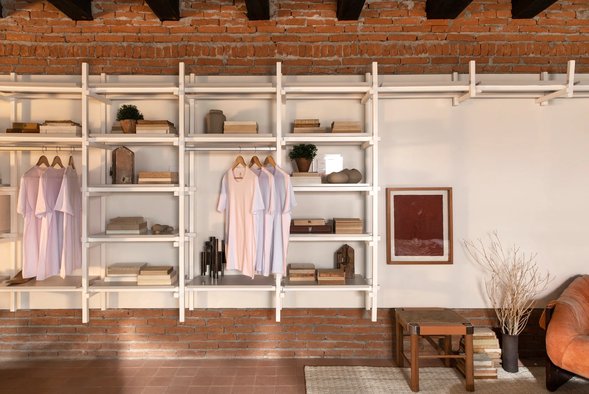 A white modular metal closet system with hanging clothes and books is mounted on a red exposed brick wall above terracotta floor tiles.