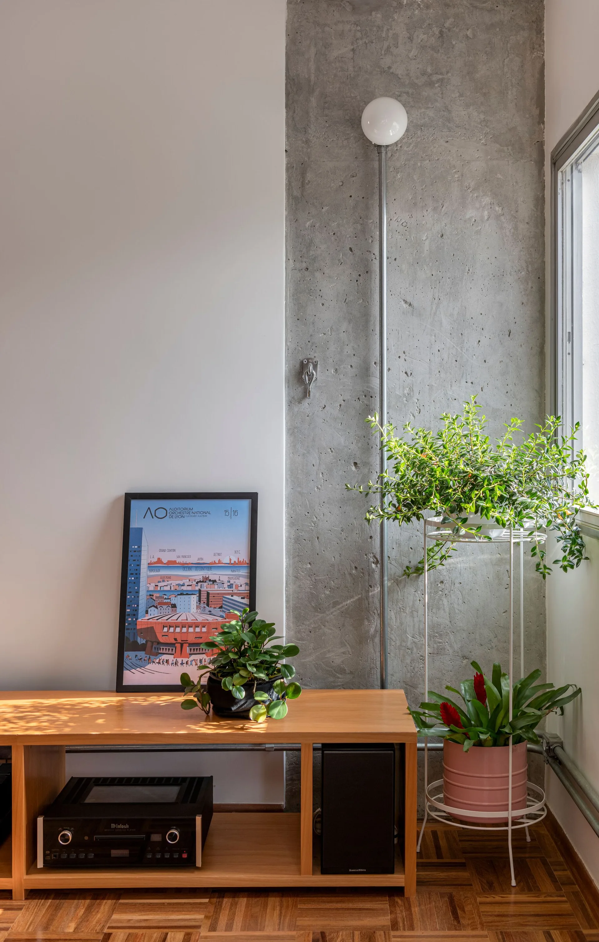 A wooden console table sits on parquet floors next to a tall concrete column, decorated with plants, a framed print, and audio equipment.