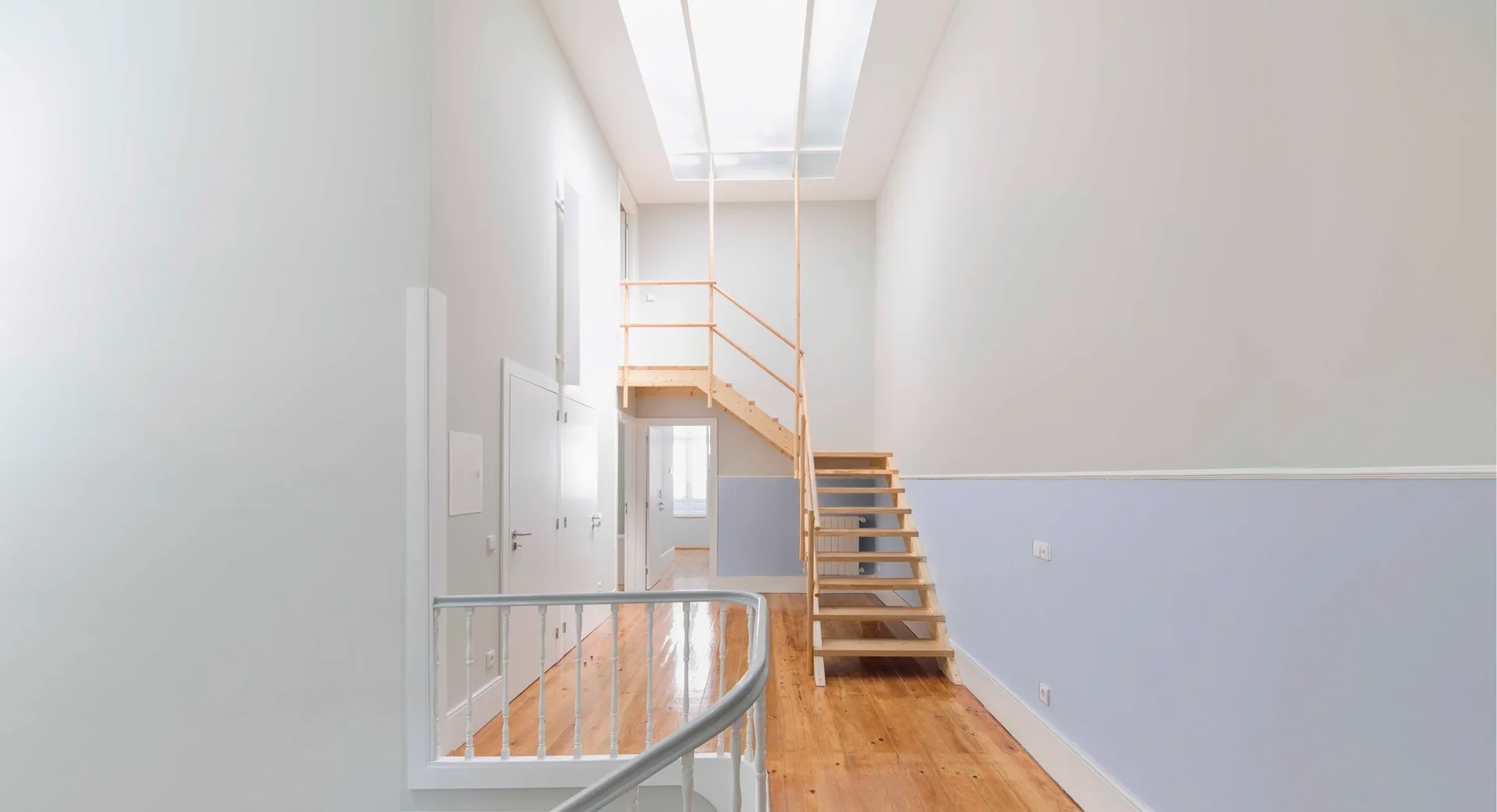 A bright hallway features an open-tread wood staircase leading upward under a large skylight, with glossy wood floors and two-tone walls.