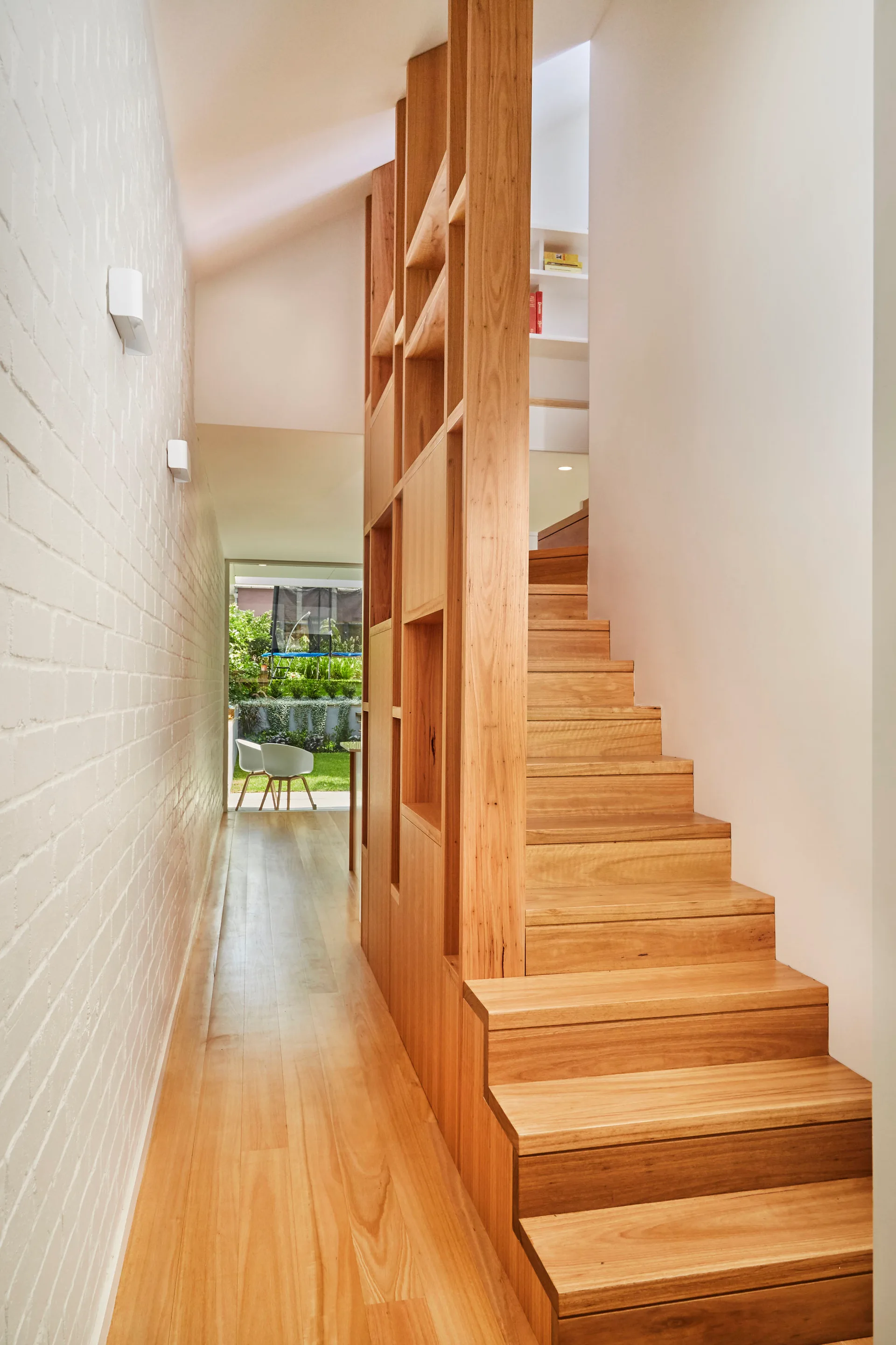 A light wood staircase with built-in shelving stands opposite a white painted brick wall in a narrow hallway leading to a garden view.