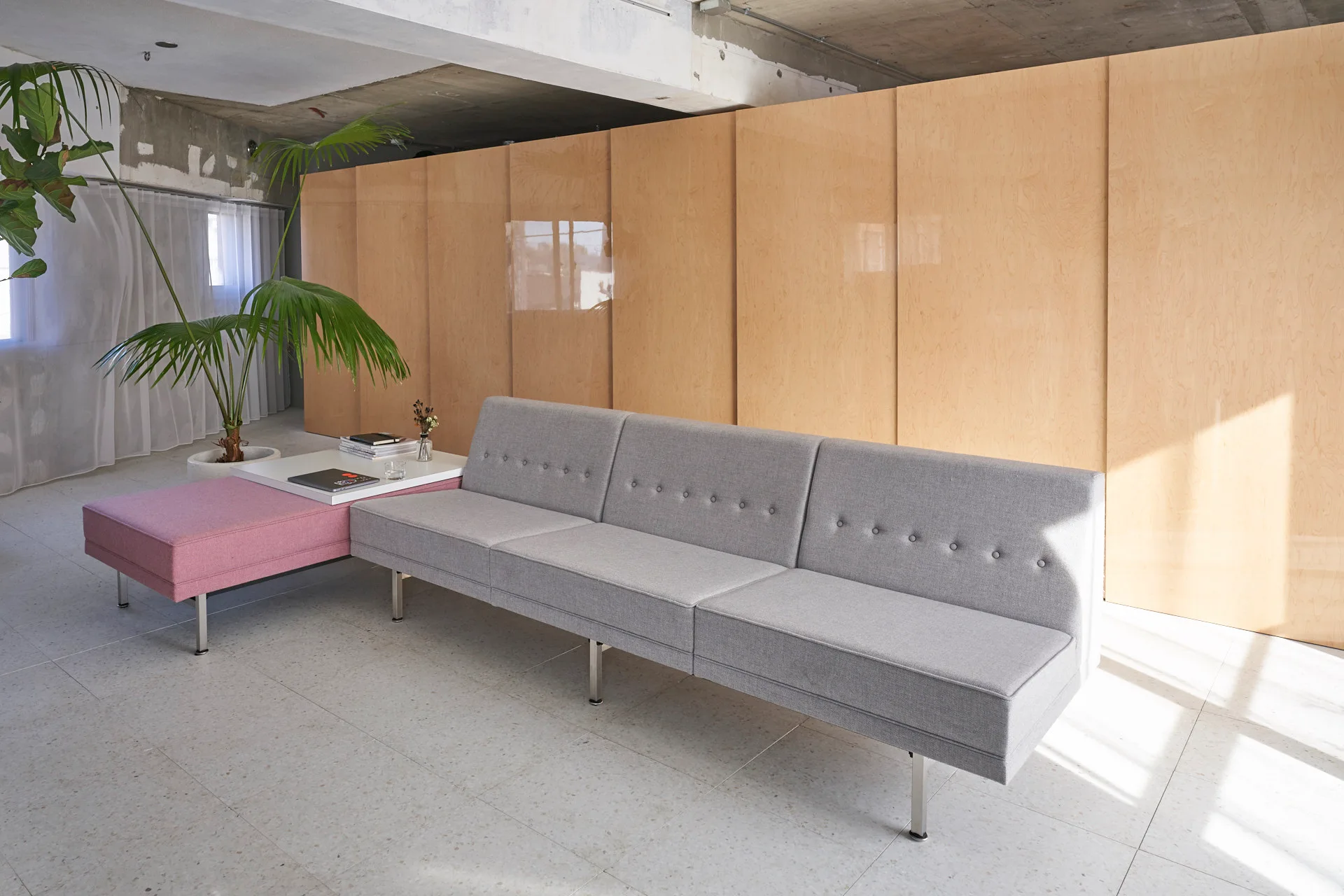 A grey button-tufted sofa sits next to a low pink bench and white side table against a backdrop of glossy wood paneling.