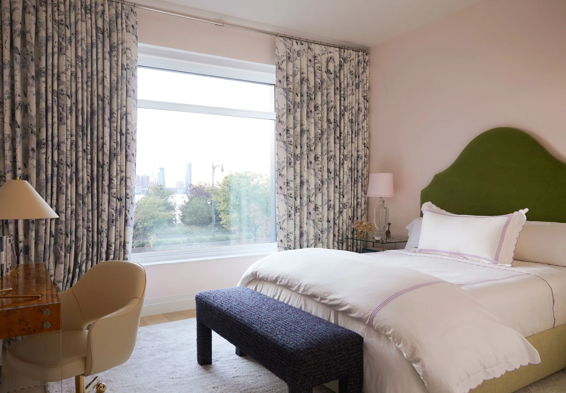 A bedroom featuring a curved green headboard on a bed with white linens, a textured purple bench, and patterned curtains framing a large window.