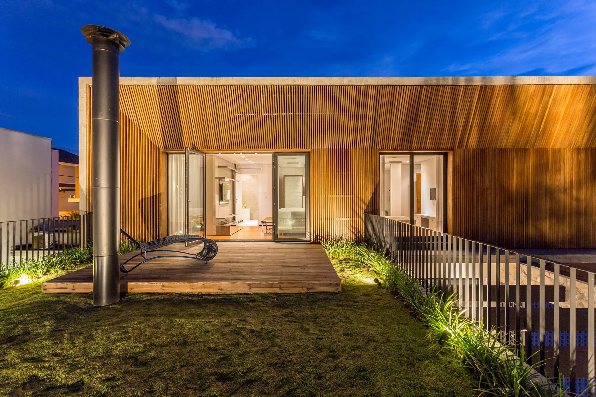 A contemporary outdoor terrace at night features a slatted wood facade, wooden deck with a lounge chair, and a grassy lawn.