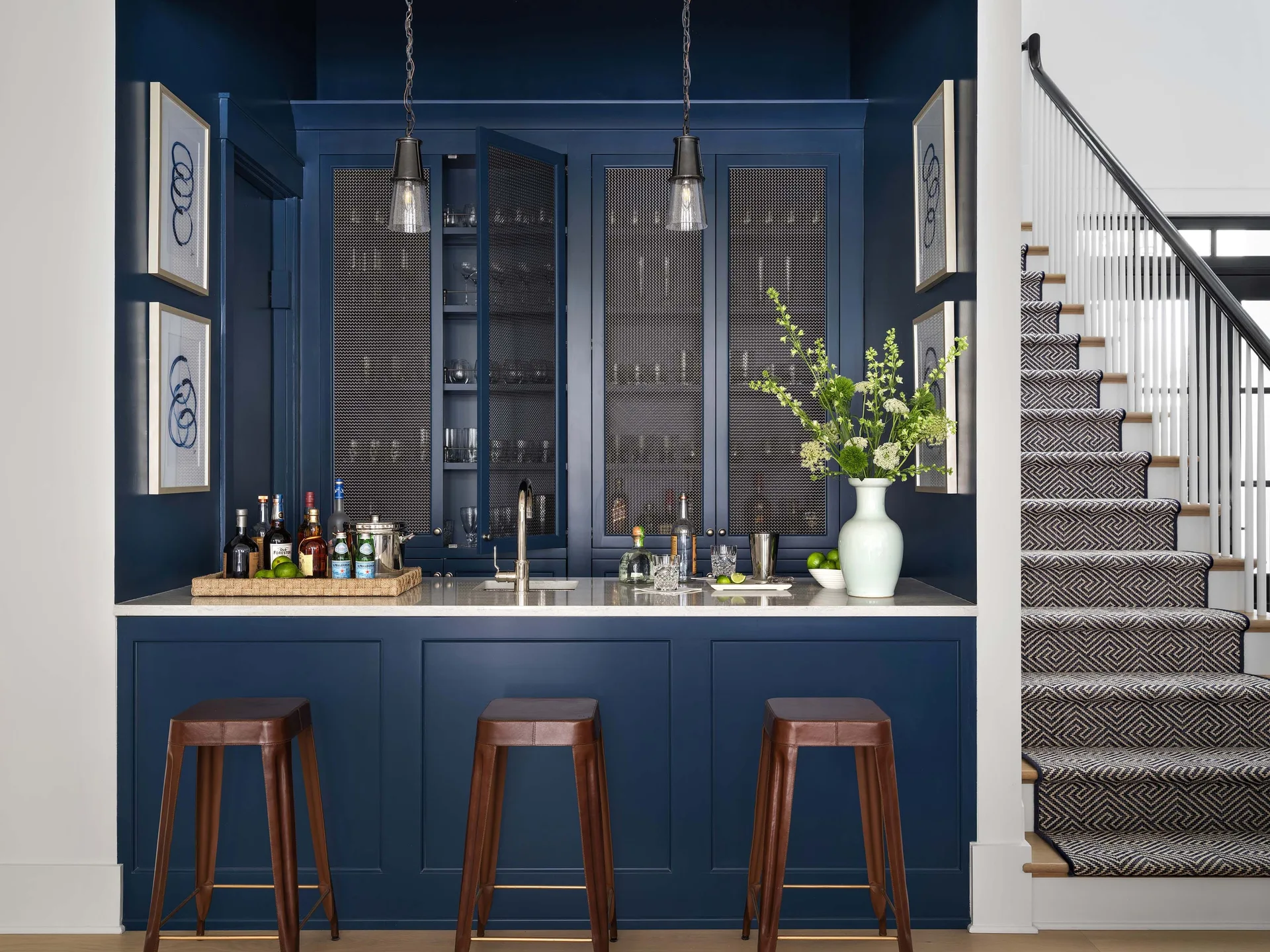 A deep navy blue home bar features wire mesh cabinet doors, three brown leather bar stools, and an adjacent staircase with a patterned runner.
