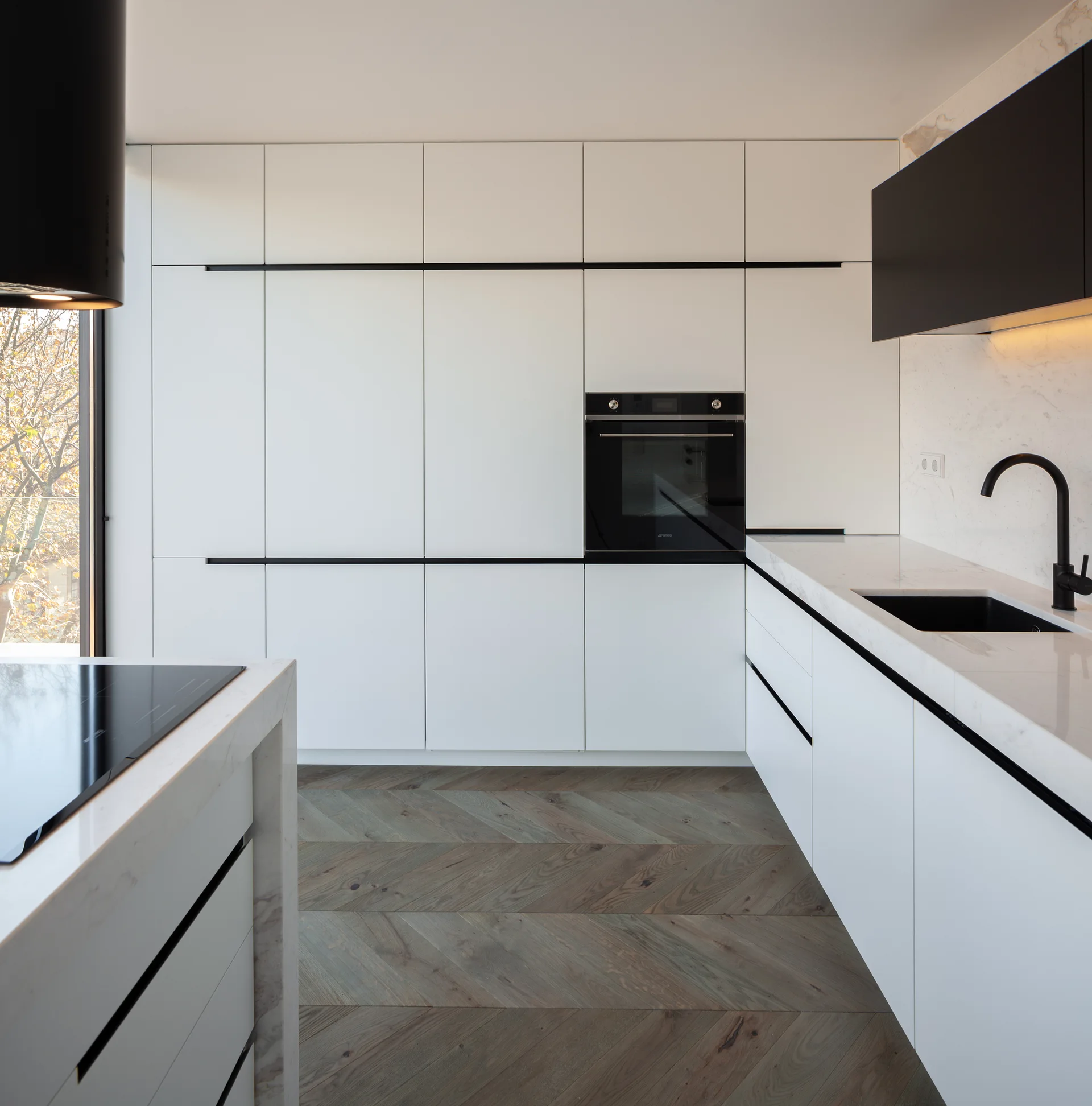 Modern kitchen with minimalist white handleless cabinets, a black built-in oven, black faucet, and herringbone pattern wood flooring.