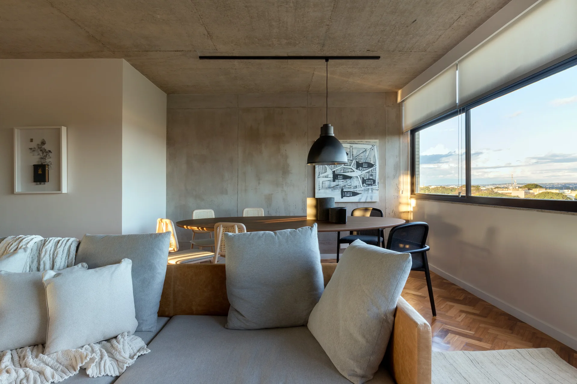 An open-plan living and dining room features an exposed concrete wall and ceiling, parquet flooring, and an oval wooden table.