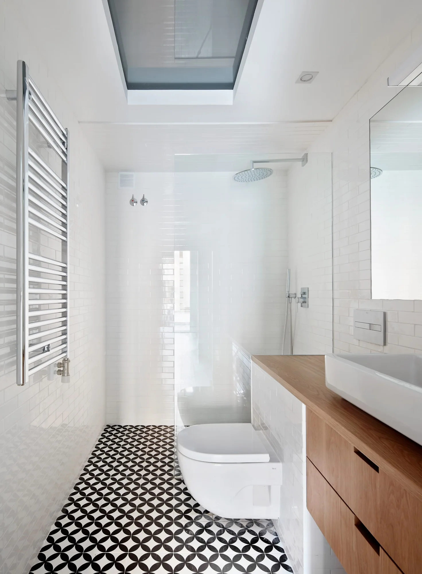 A modern, narrow bathroom with white subway tile walls, a skylight, a wood vanity, and black and white geometric floor tiles.
