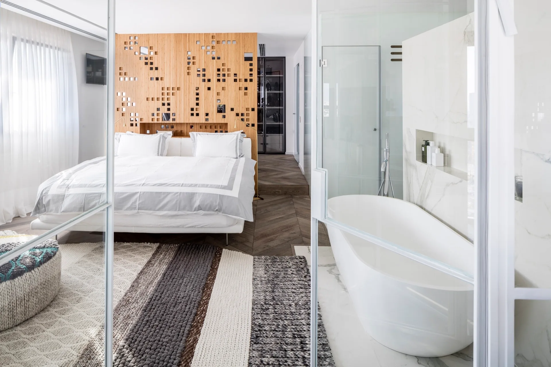 A contemporary bedroom featuring a glass partition that opens to an ensuite bathroom with a freestanding white tub and marble wall.
