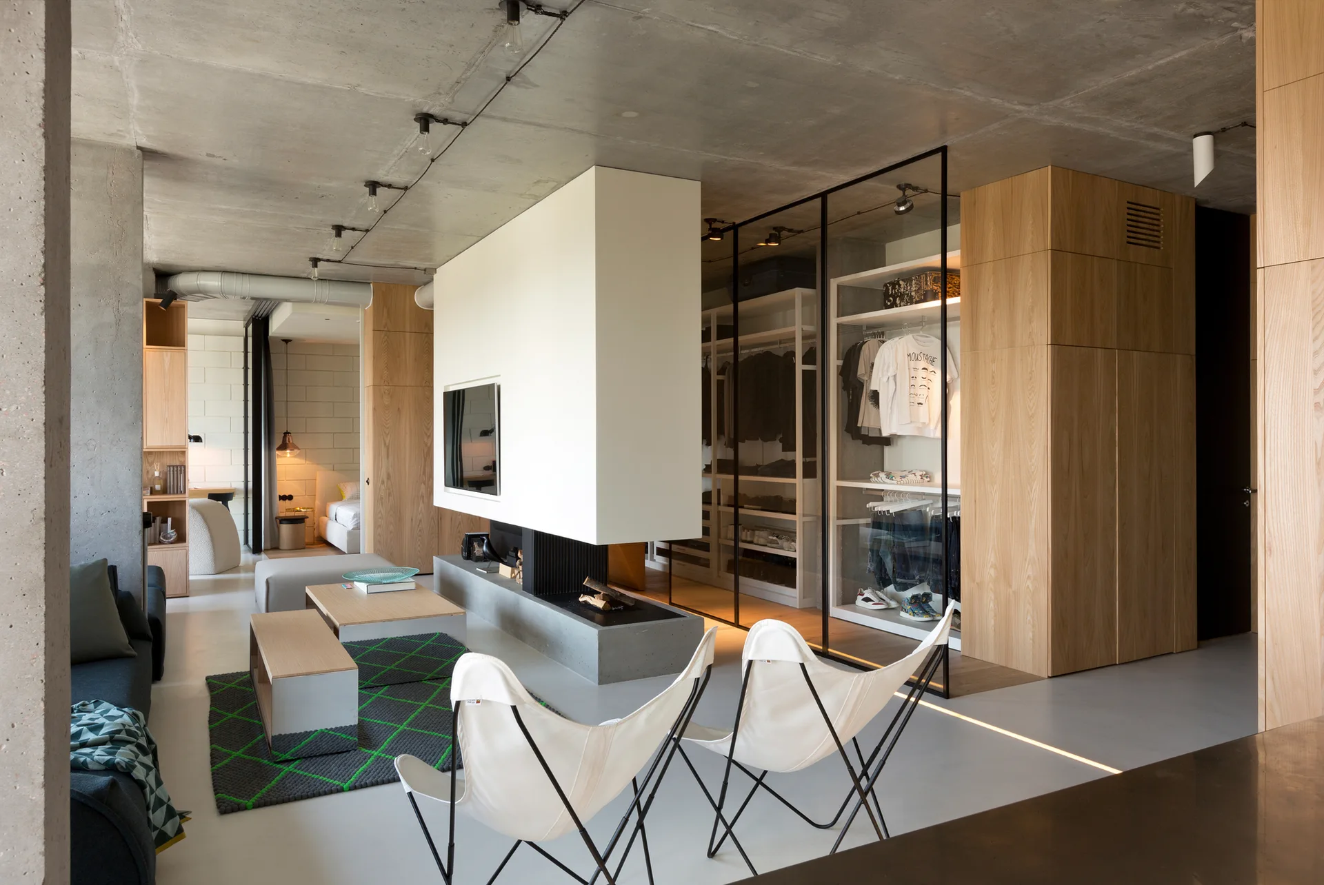 An open-concept living area features an exposed concrete ceiling, a central white fireplace partition, and a glass-enclosed wardrobe.