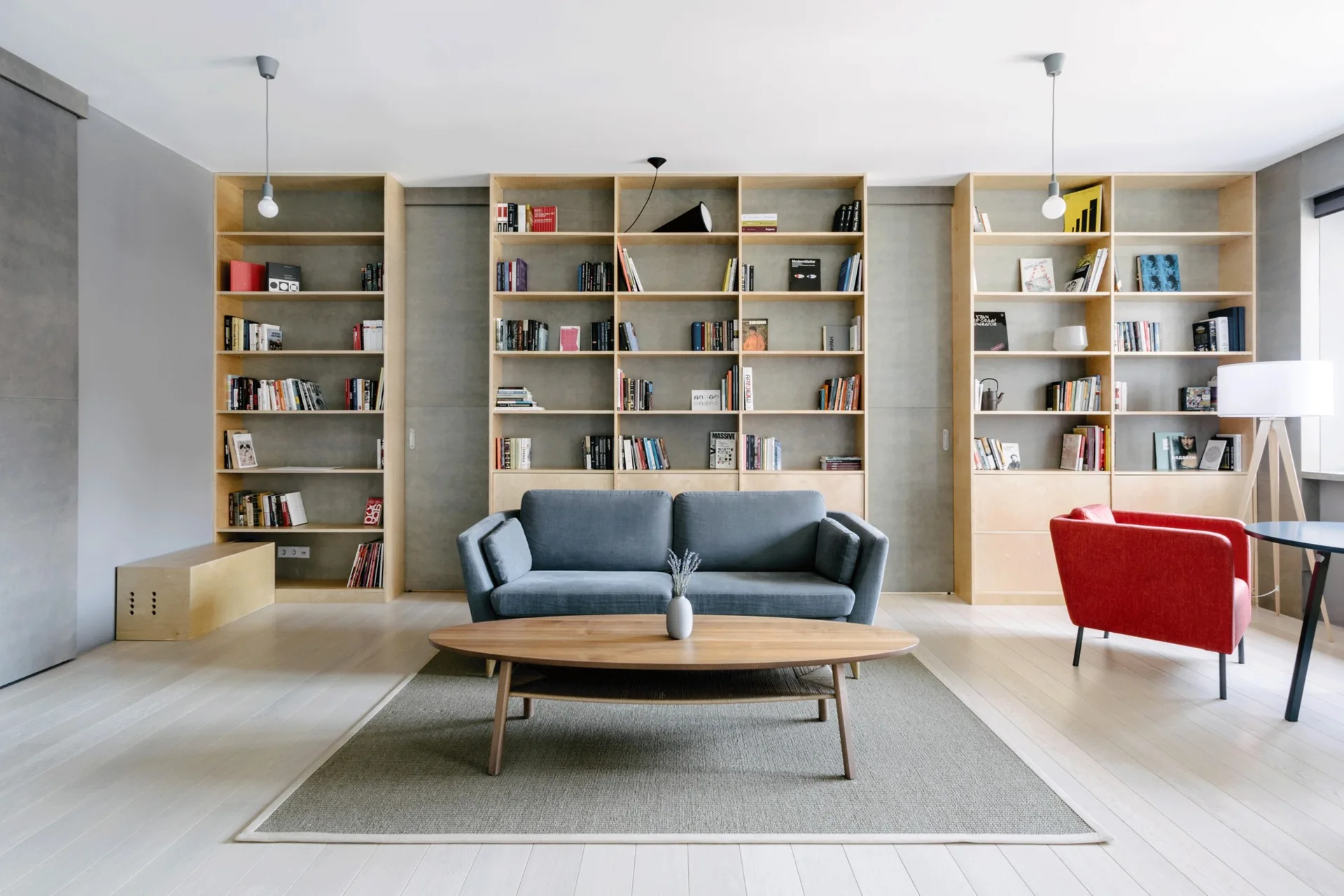 A modern living room features a blue sofa, an oval wood coffee table, a red armchair, and floor-to-ceiling light wood bookshelves.