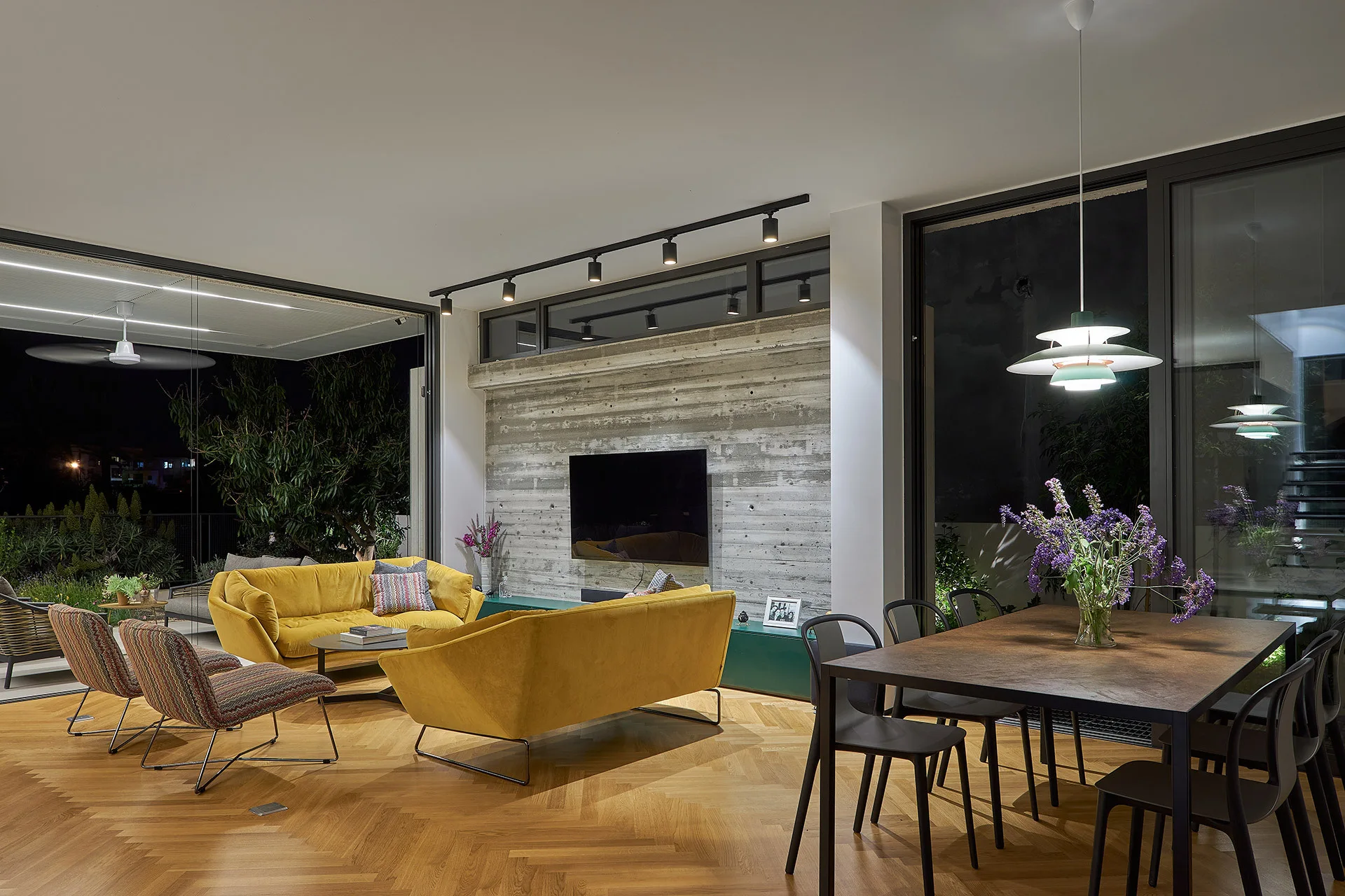 An open living room features two yellow velvet sofas, an exposed concrete wall with a TV, herringbone wood flooring, and a nearby dining set.