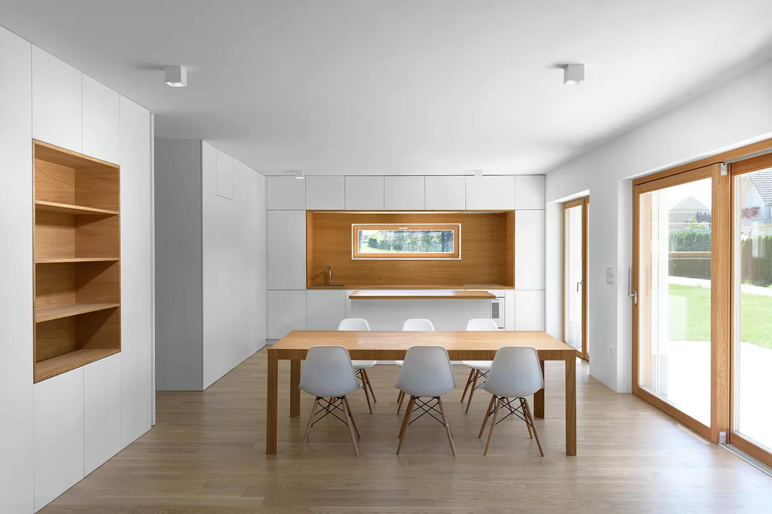 A light wood dining table with six white chairs stands in front of white minimalist kitchen cabinets featuring a warm wood backsplash niche.