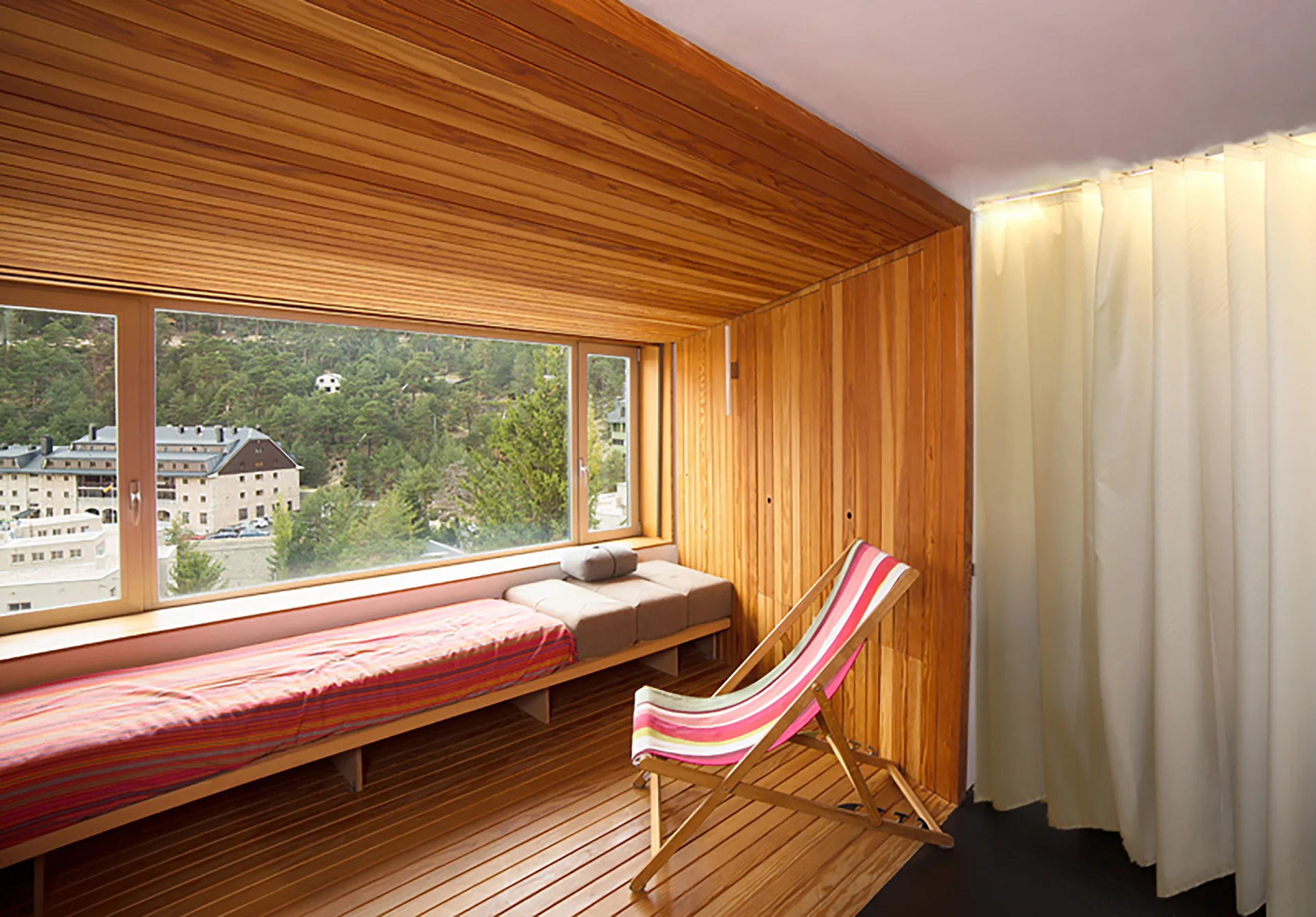 A wood-paneled bedroom alcove features a built-in window seat looking out at mountains, alongside a striped deck chair and cream curtains.