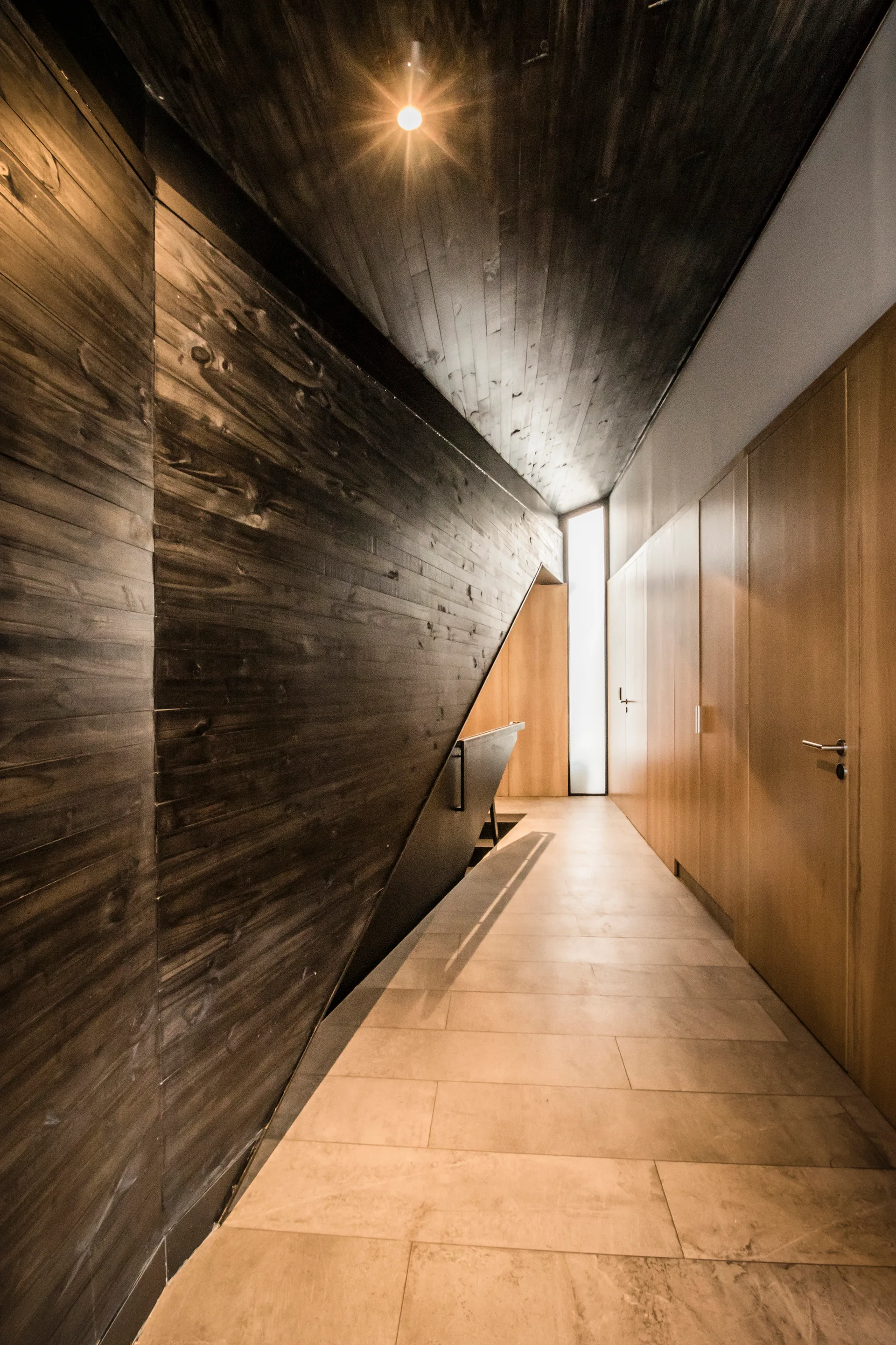 A modern hallway features dark wood paneled walls, flush light wood closet doors, and beige stone floor tiles.