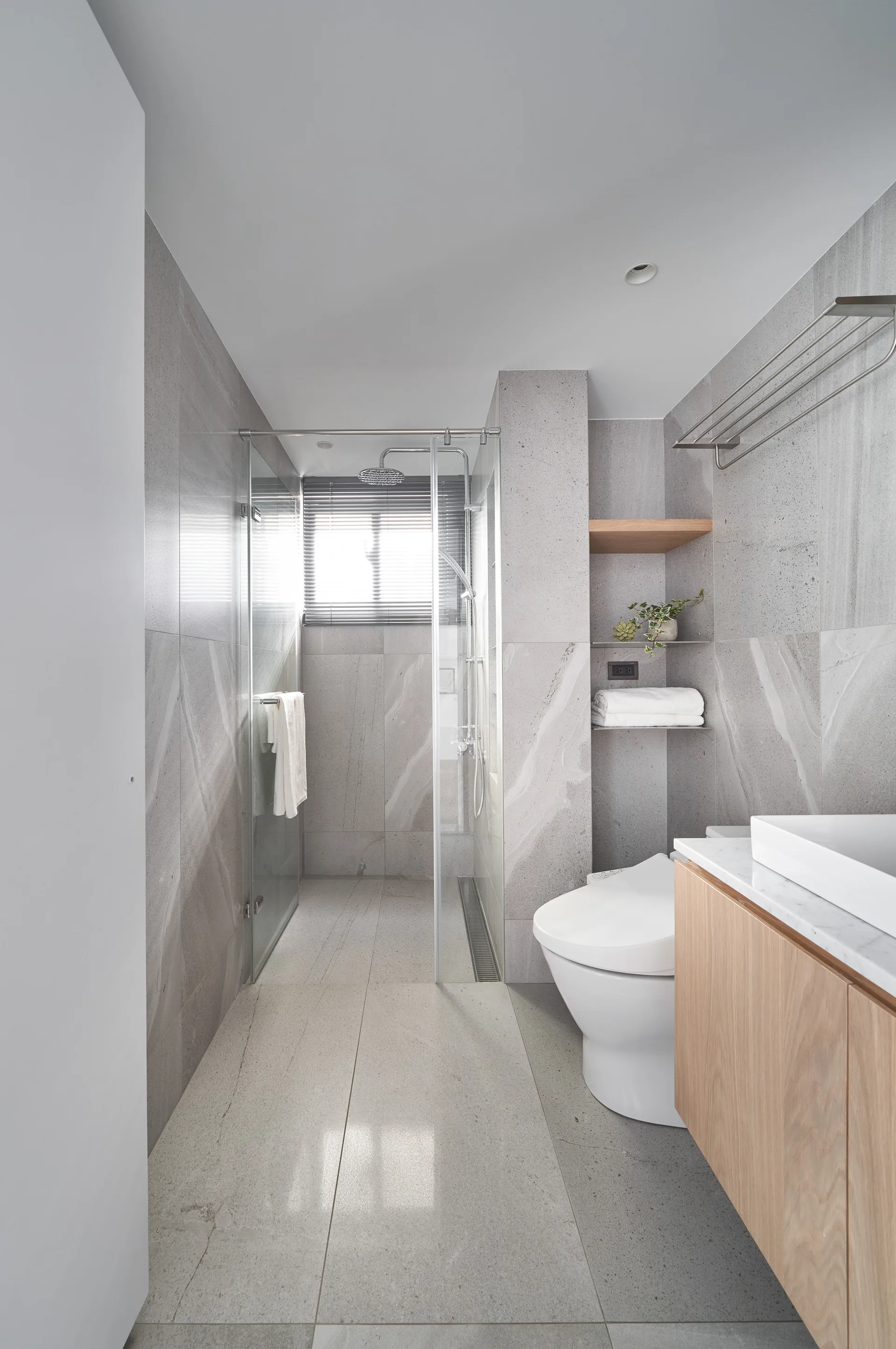 A contemporary grey bathroom featuring a walk-in glass shower, a floating wooden vanity, a white toilet, and recessed wall shelving.