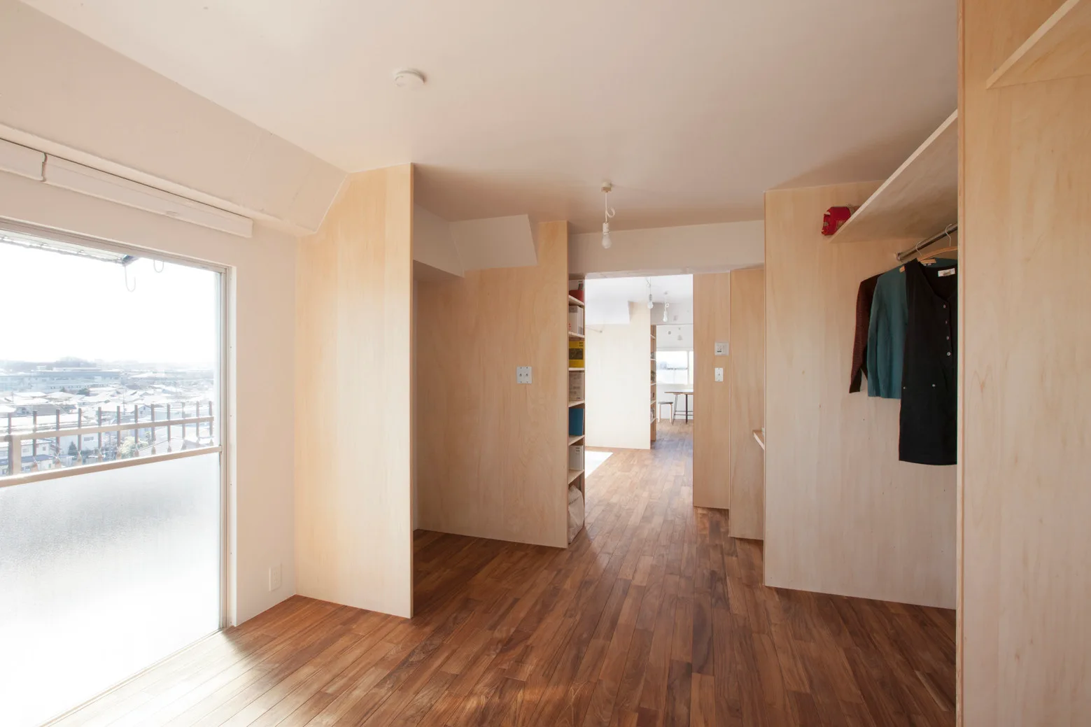 A minimalist interior space features light plywood partition walls, medium-brown wood plank flooring, a large window, and built-in open closet storage.