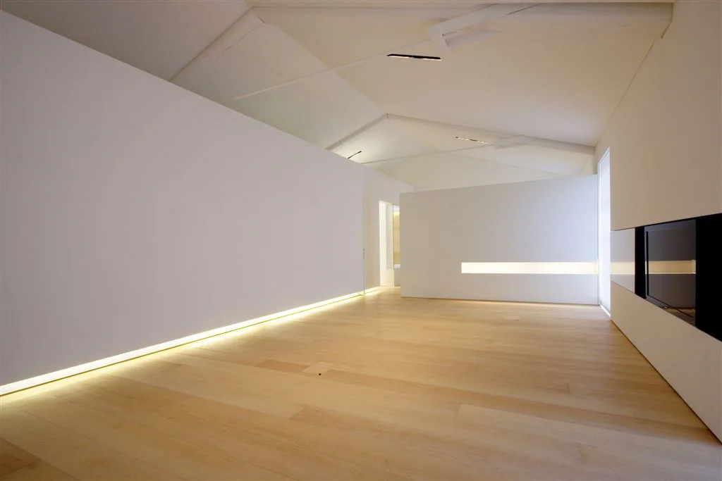 A minimalist living space featuring a vaulted ceiling, light wood floors, smooth white walls, and built-in floor-level cove lighting.