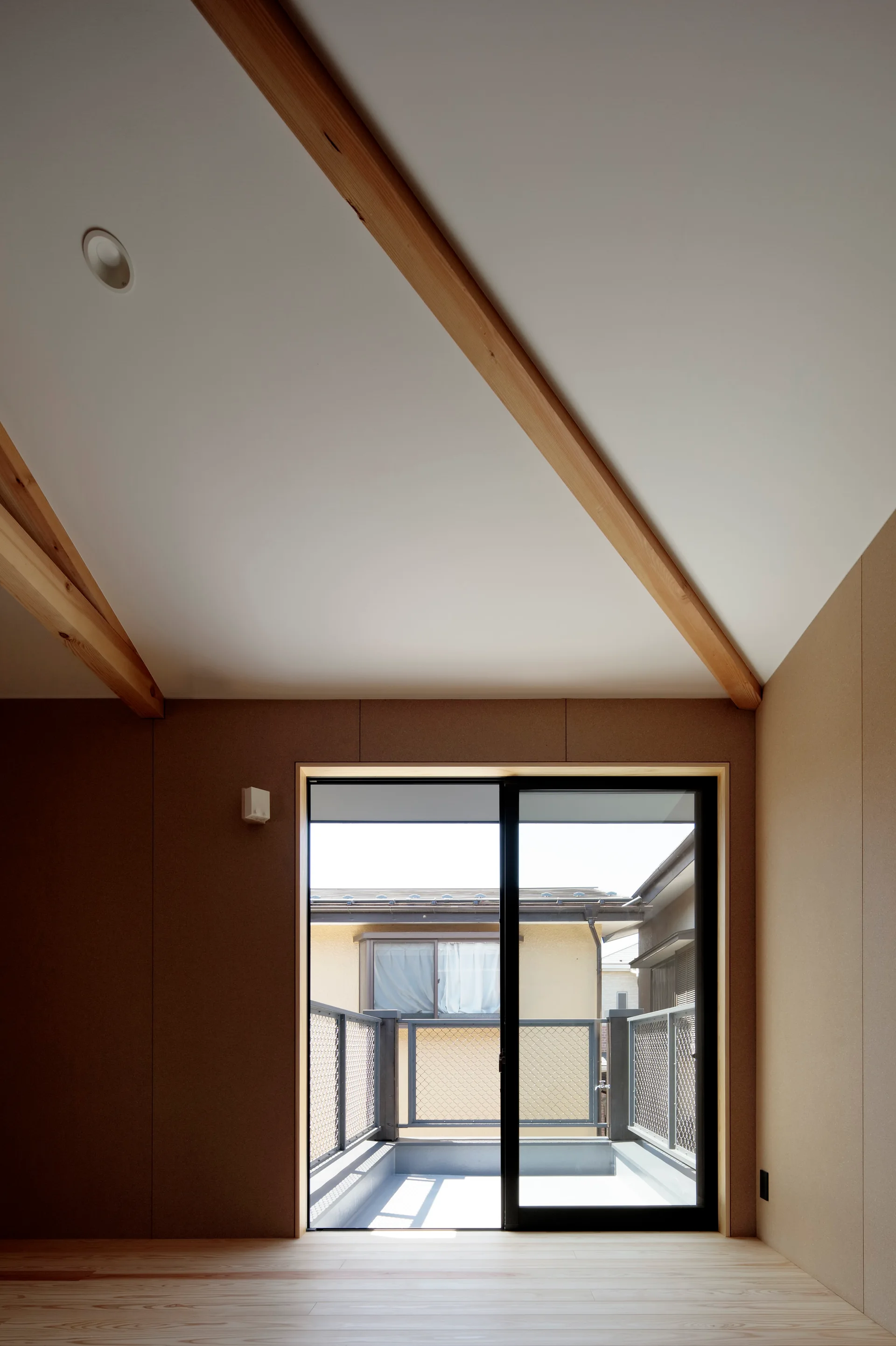 An empty room with tan walls features light wood flooring, exposed diagonal ceiling beams, and a sliding glass door opening to a balcony.